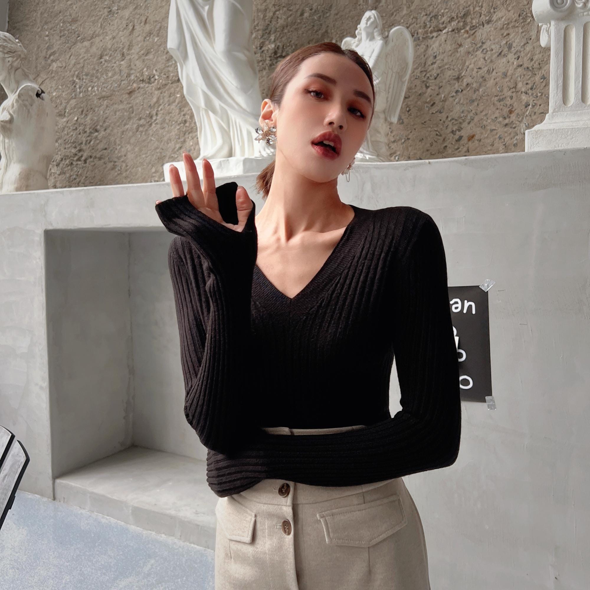 A woman wearing a black sweater and beige pants stands in front of a wall with statues, making a hand gesture.