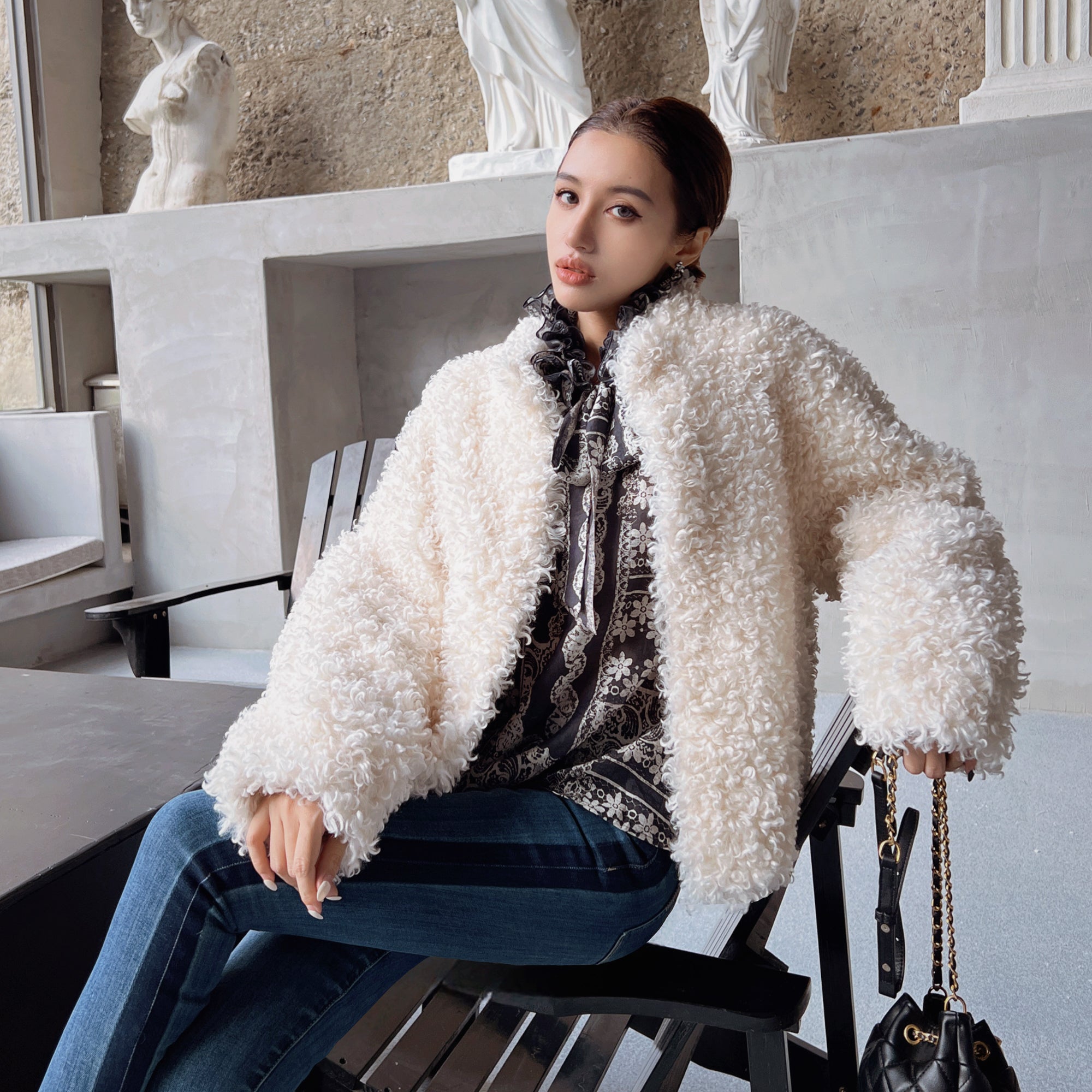 A woman wearing a white fluffy coat and holding a black purse is sitting on a bench in an indoor setting with a stone wall and statues in the background.