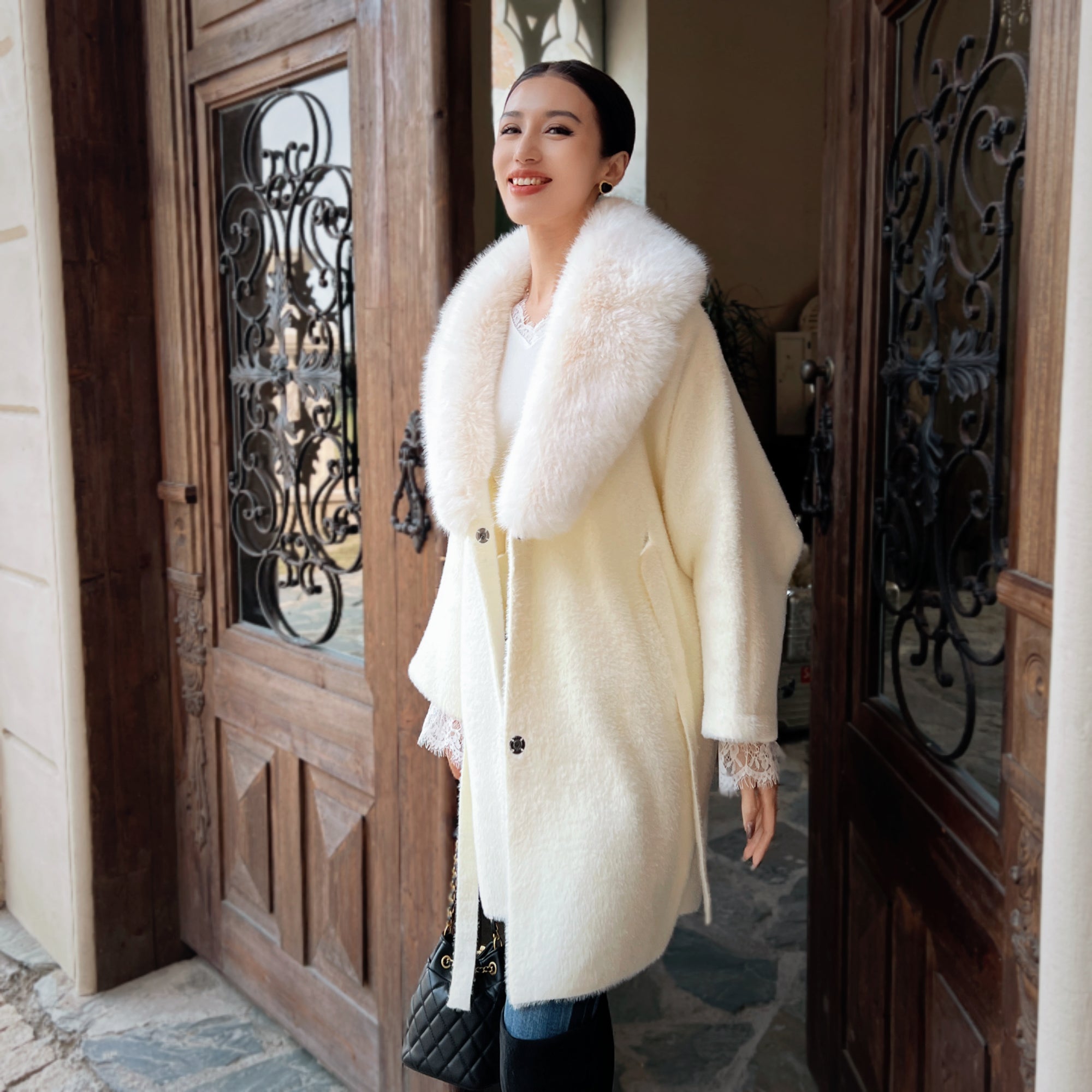 A woman wearing a white fur coat and smiling at the camera, standing in front of a wooden door with wrought iron details.