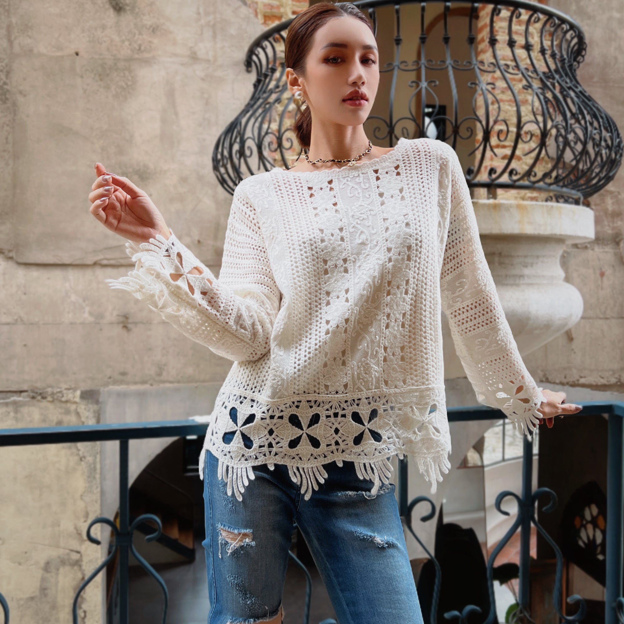 A woman wearing a white lace top with fringed edges and distressed jeans stands in front of a wrought iron balcony.