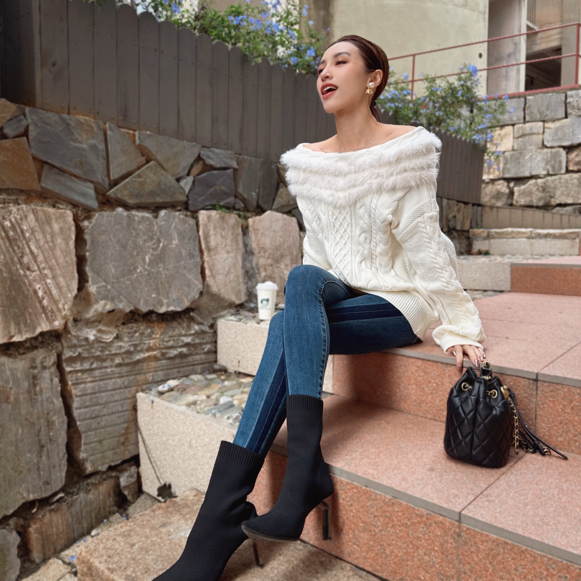 A woman in a white sweater and blue jeans sits on a set of stairs, holding a black purse, with a stone wall and flowers in the background.