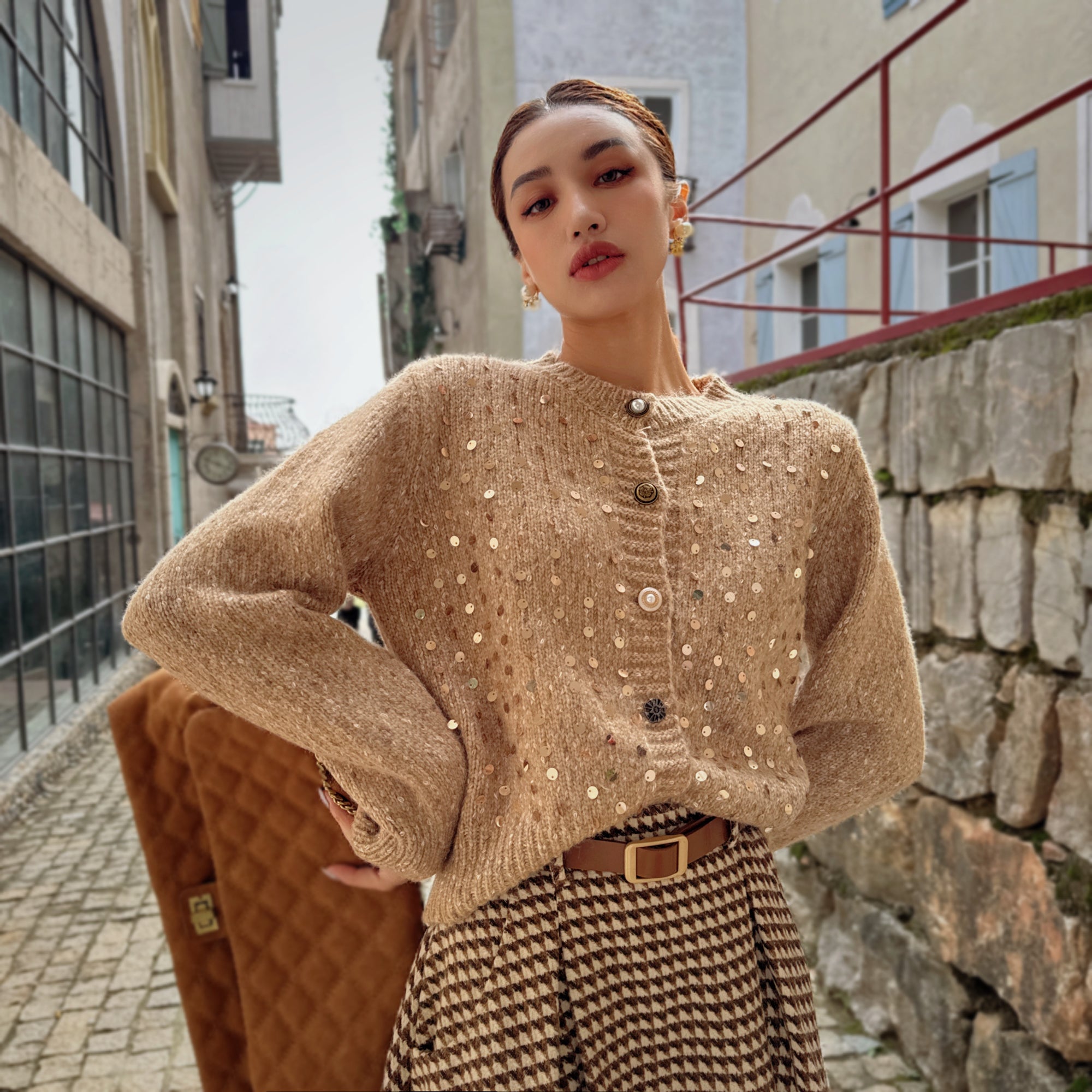 A woman wearing a beige sweater with white polka dots and a brown skirt stands in front of a stone wall.