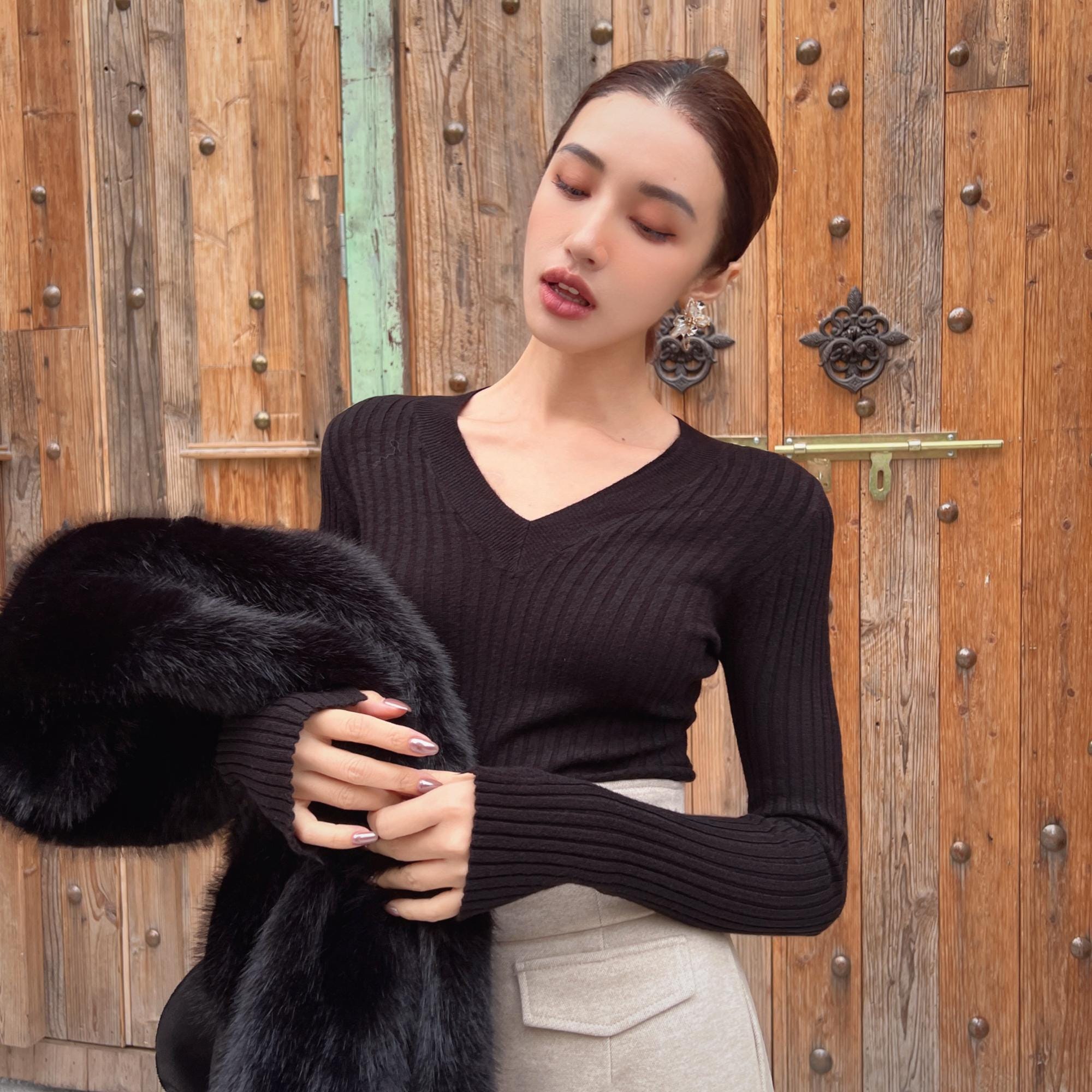 A woman in a black sweater and skirt stands in front of a wooden door, holding a black fur coat.