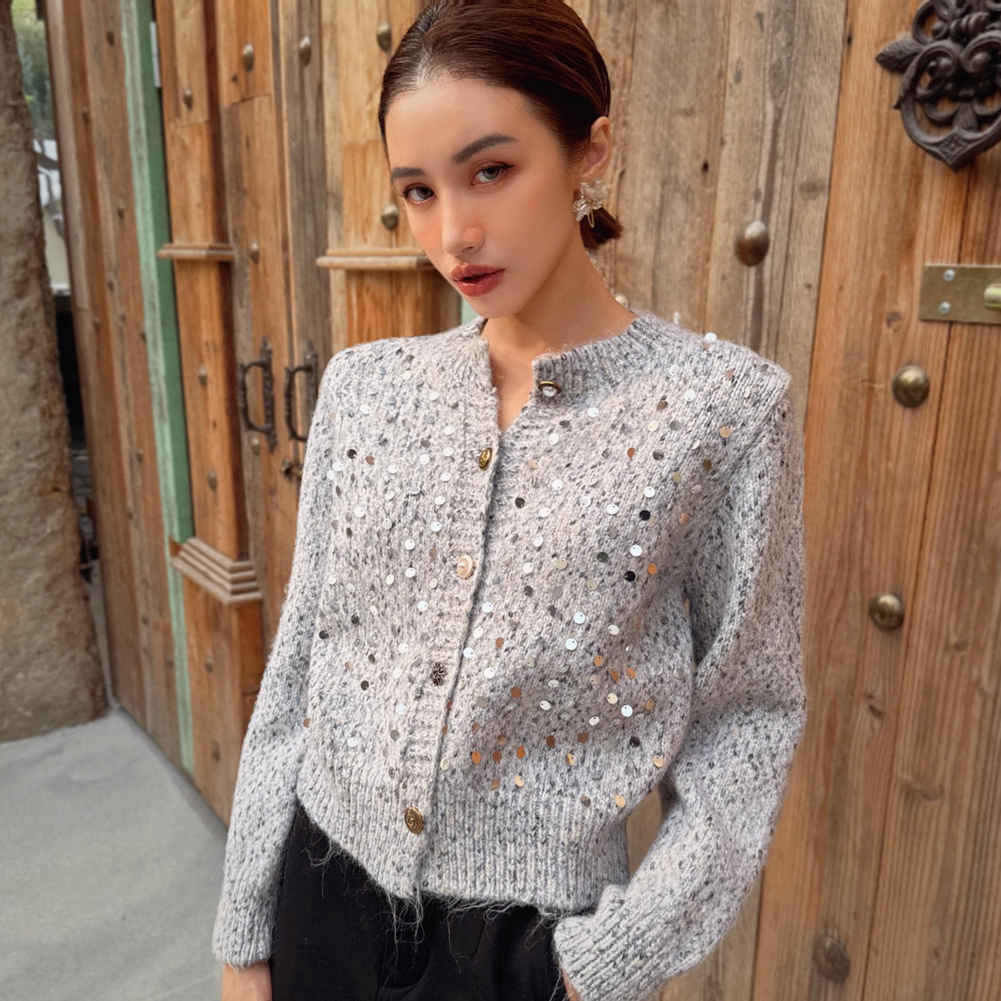 A young woman wearing a gray sweater with silver sequins stands in front of a wooden door.