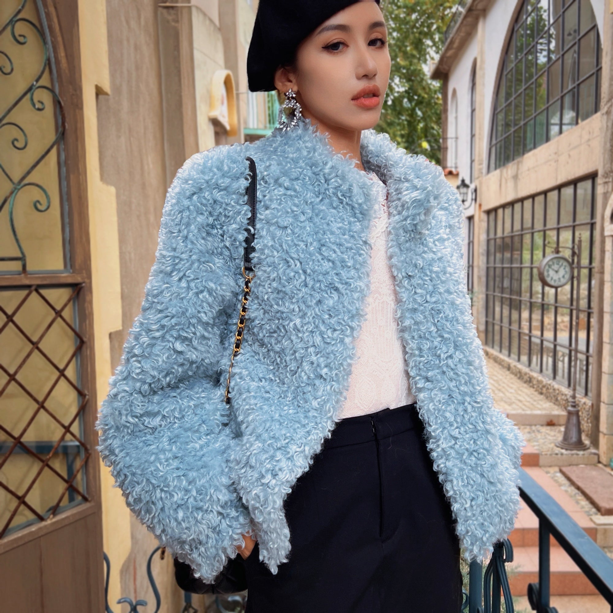 A woman wearing a light blue coat, black pants, and a black hat stands in front of a building with a wrought iron gate.