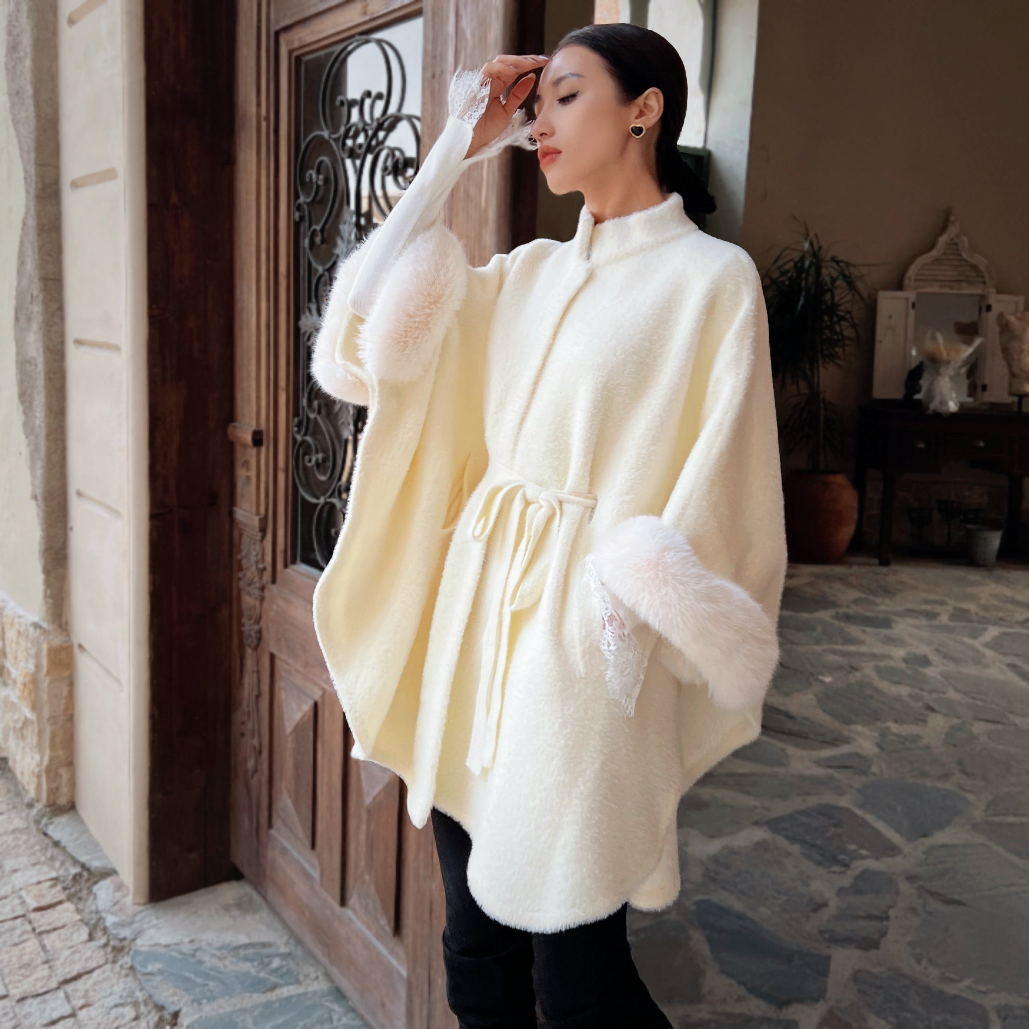 A woman wearing a white fur-trimmed poncho stands in front of a wooden door, with a potted plant visible in the background.