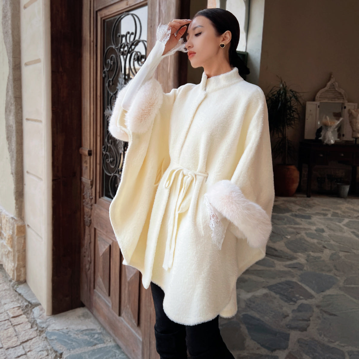 A woman wearing a white fur-trimmed poncho stands in front of a wooden door, with a potted plant visible in the background.