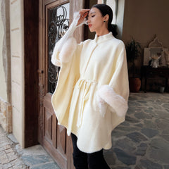 A woman wearing a white fur-trimmed poncho stands in front of a wooden door, with a potted plant visible in the background.