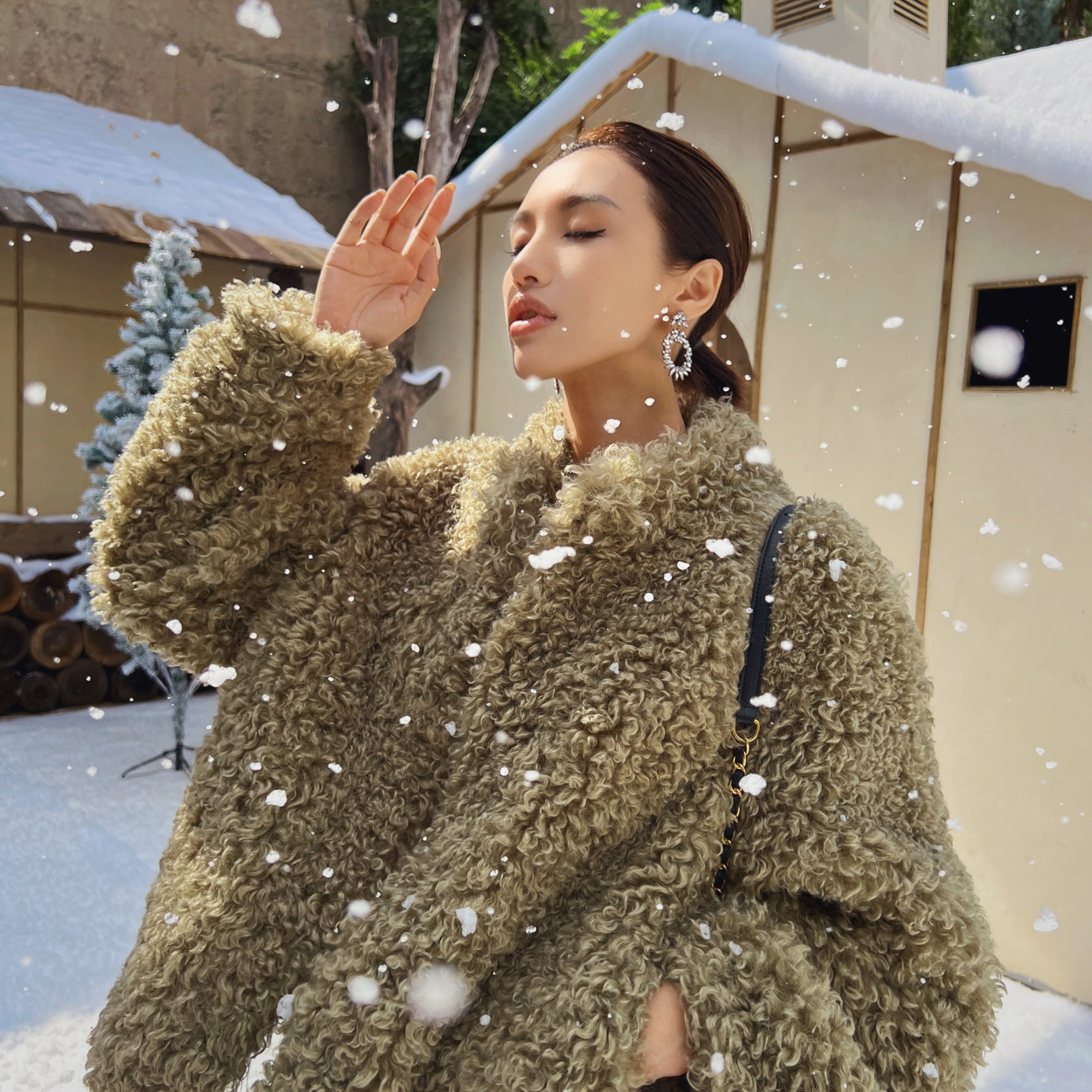 A woman in a brown furry coat stands in the snow, her hand raised to her face as snowflakes fall around her.