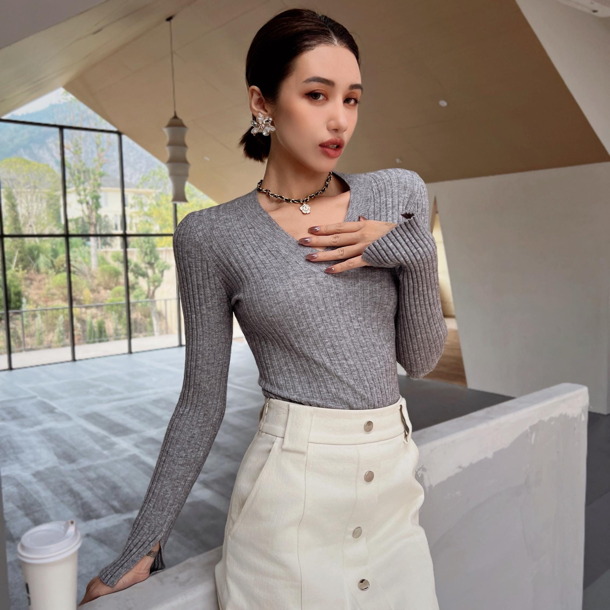 A woman wearing a gray sweater and white skirt stands in a room with large windows, holding a cup of coffee.