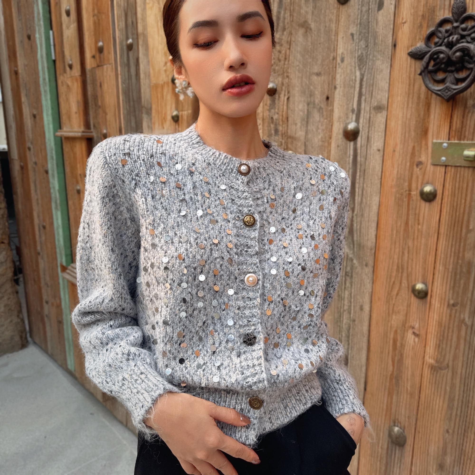A woman wearing a gray sweater with silver buttons and a patterned design stands in front of a wooden door.