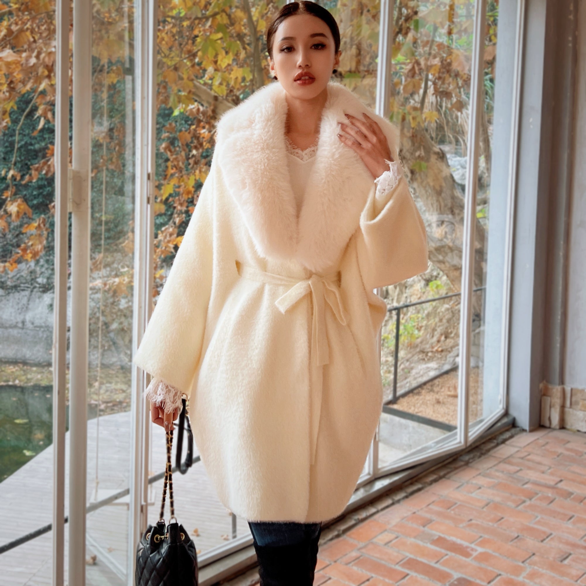A woman wearing a long, white fur coat with a fur collar stands in front of a window, holding a black purse.