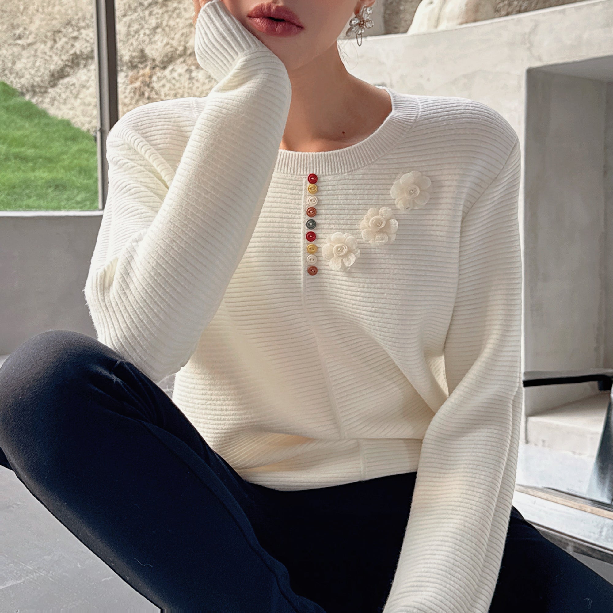 A woman wearing a white sweater with colorful buttons and a flower design, sitting with her hand on her chin.