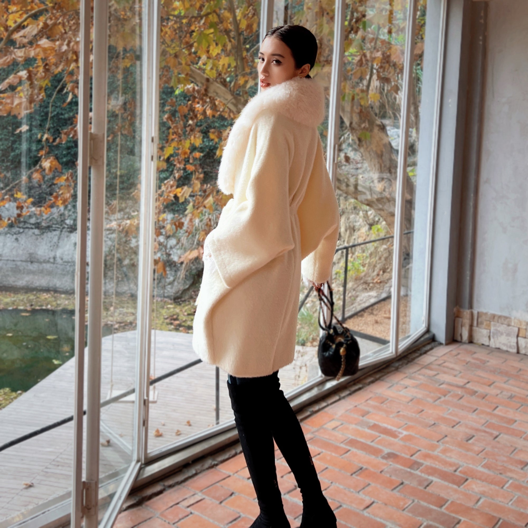 A woman in a white coat stands in front of a window, looking out at a scenic view of trees and a body of water.