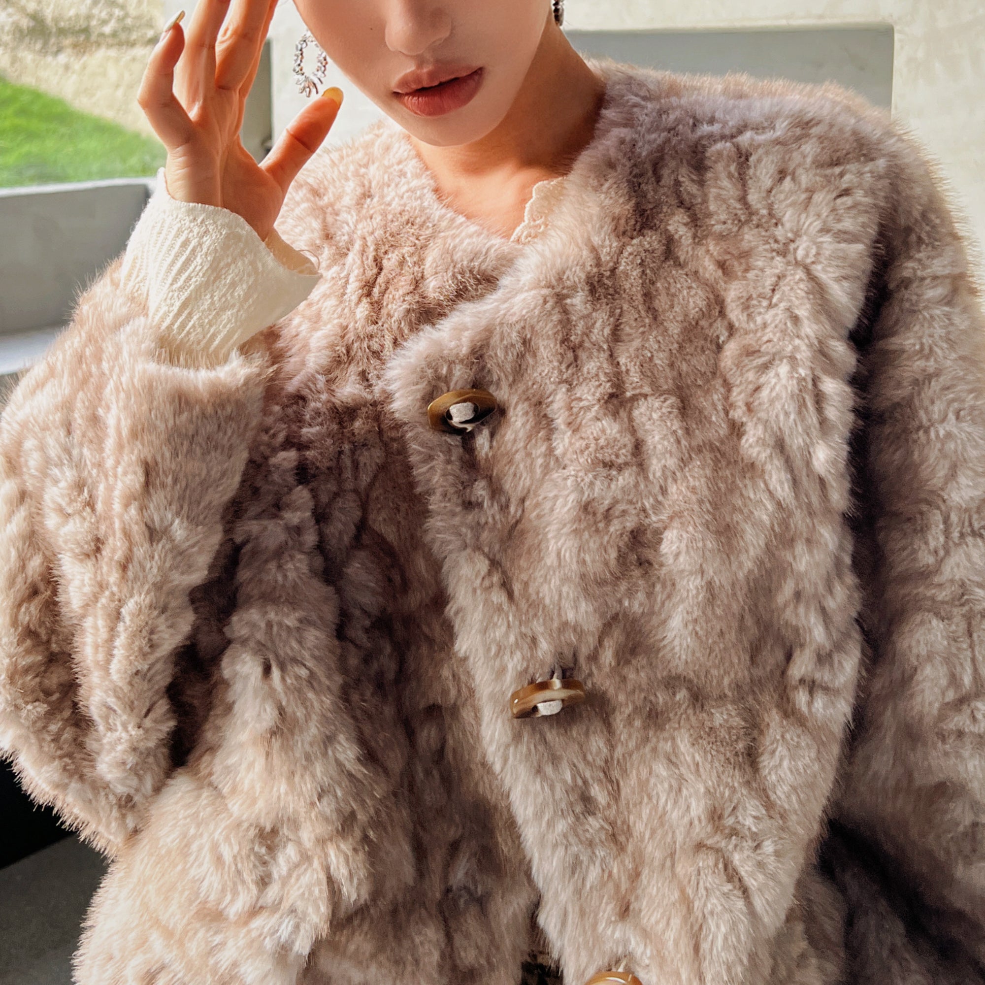 A close-up of a person wearing a beige fur coat, with their hand resting on their face.