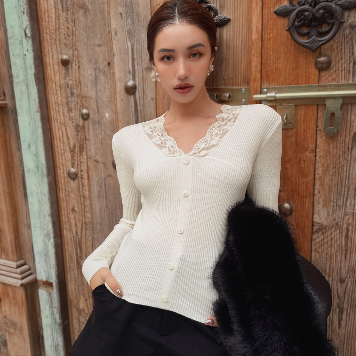 A woman wearing a white cardigan with lace trim and holding a black fur coat stands in front of a wooden door.