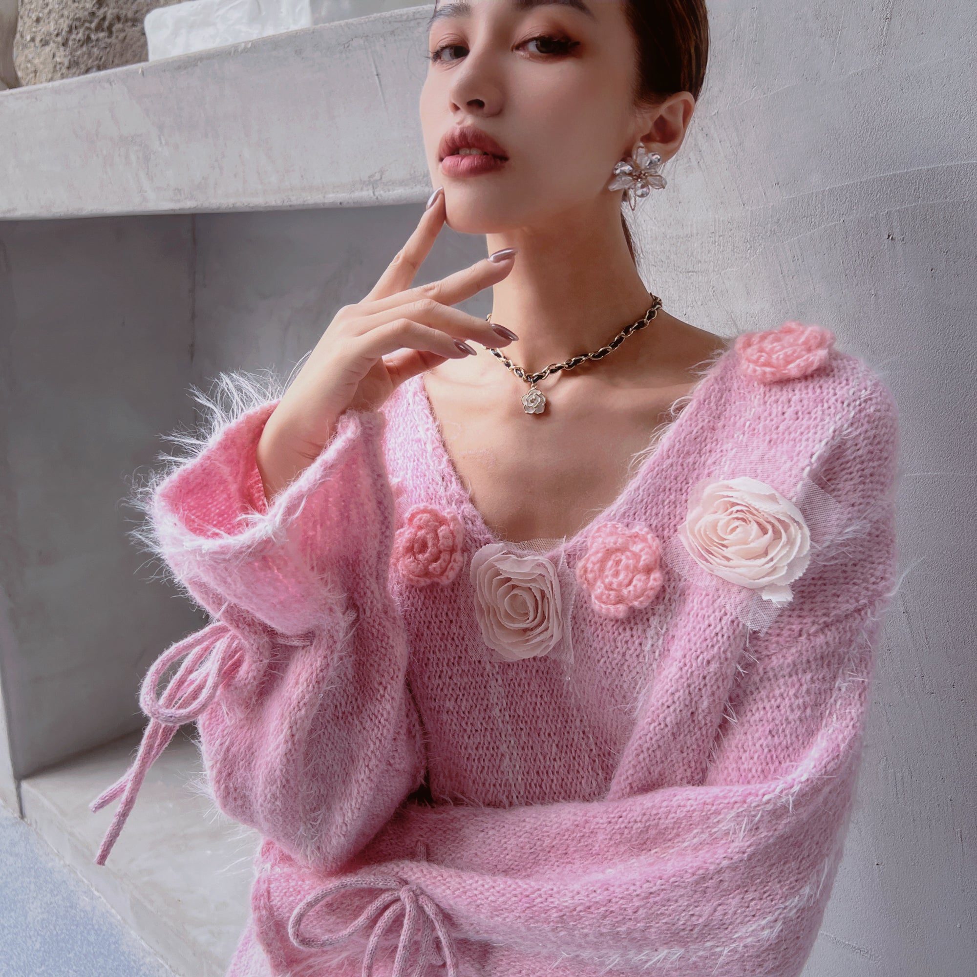 A woman wearing a pink sweater with pink flowers and a necklace is posing for a photograph.