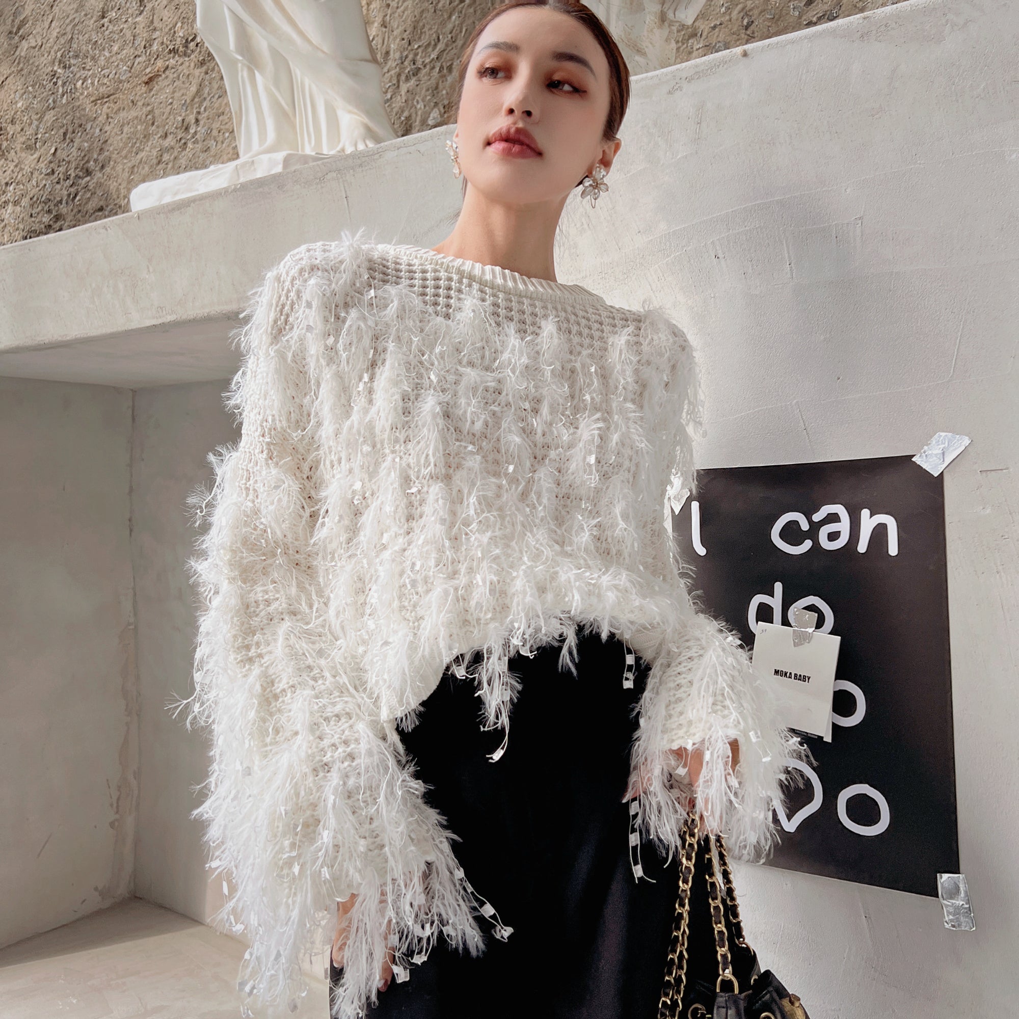 A woman wearing a white, fluffy sweater and black skirt stands in front of a black and white poster with the text "I can do anything" on it.