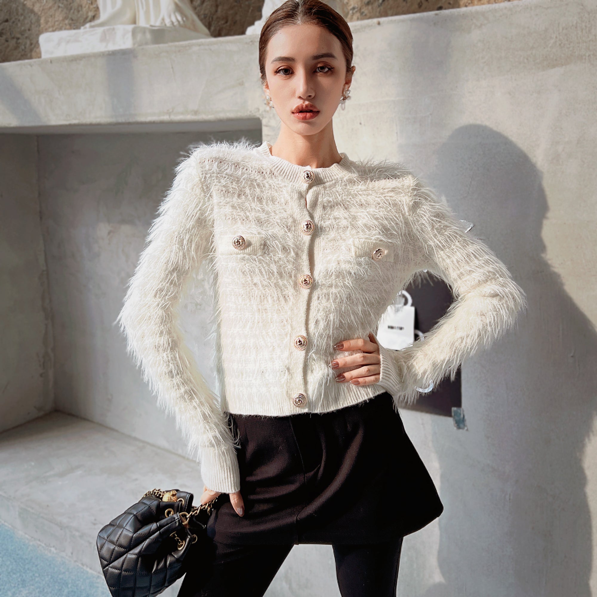 A woman wearing a white furry cardigan and black skirt, posing in front of a concrete wall. She is holding a black purse in her hand.
