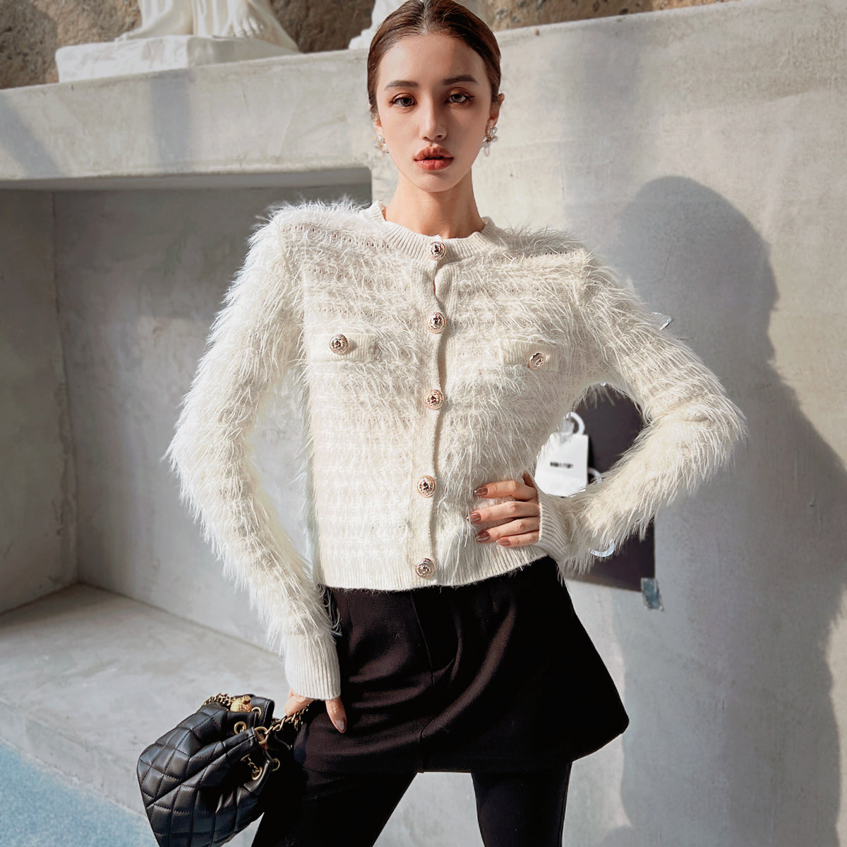 A woman wearing a white furry cardigan and black skirt, posing in front of a concrete wall. She is holding a black purse in her hand.