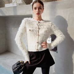 A woman wearing a white furry cardigan and black skirt, posing in front of a concrete wall. She is holding a black purse in her hand.