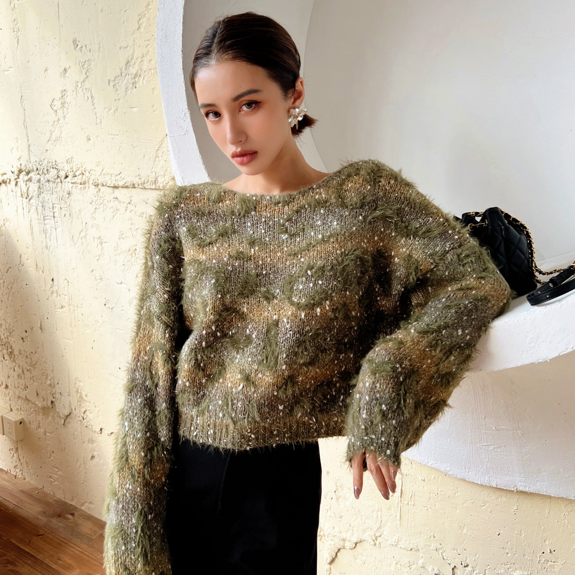 A woman wearing a green and brown fuzzy sweater stands in front of a white wall, leaning against it with her hand on her hip.