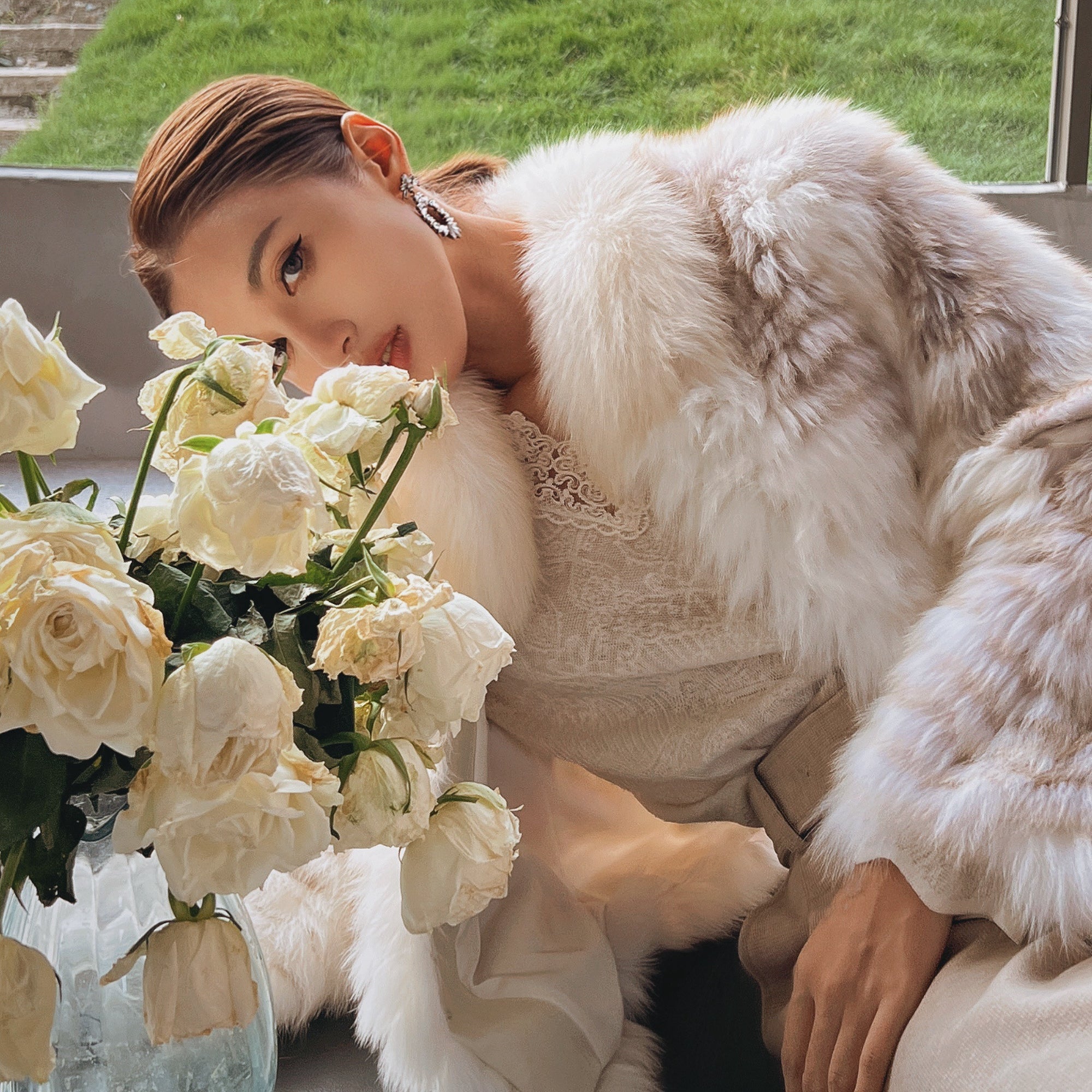 A woman in a white fur coat is leaning against a window, with a vase of white roses in front of her.