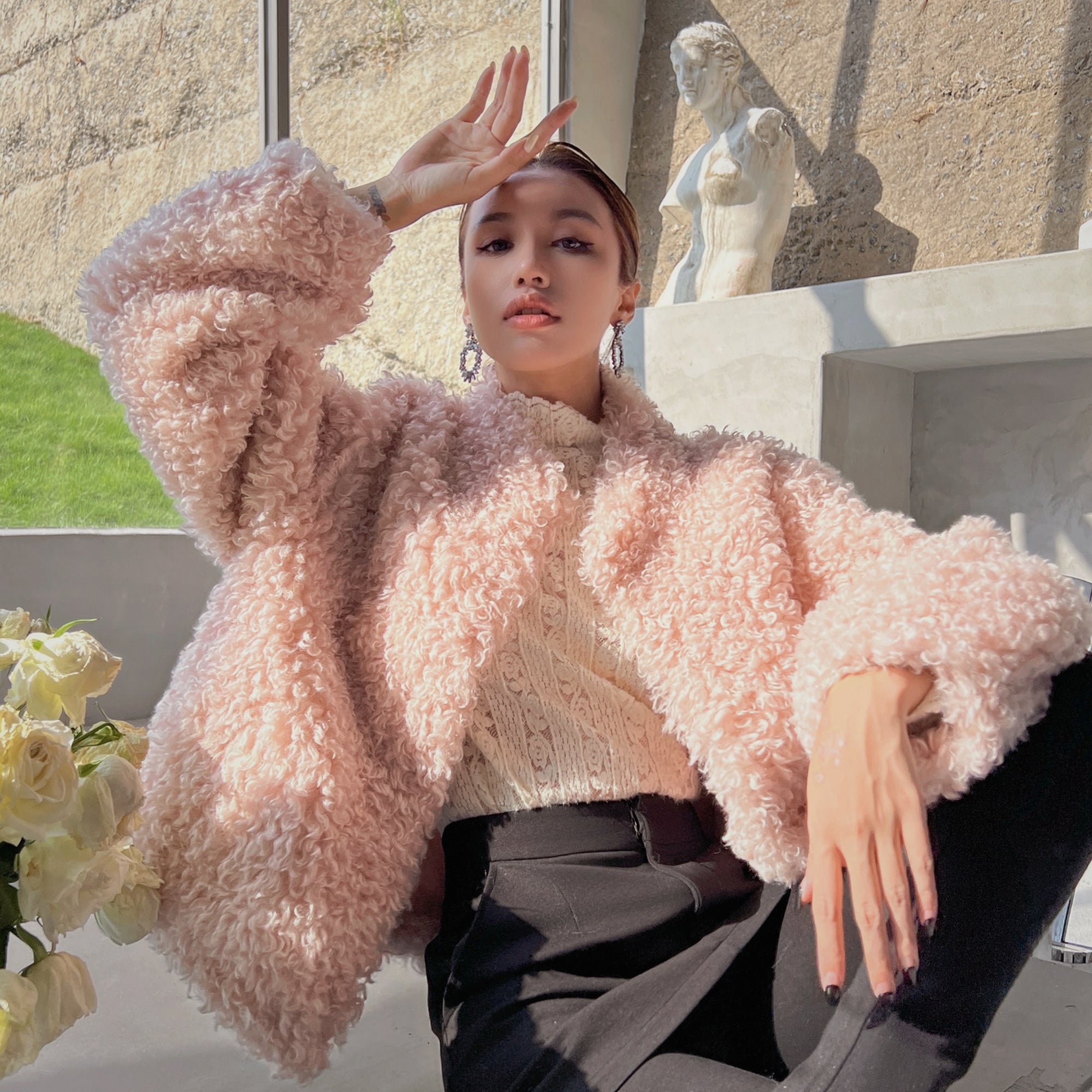 A woman wearing a pink furry coat and sitting in front of a window with a statue in the background.