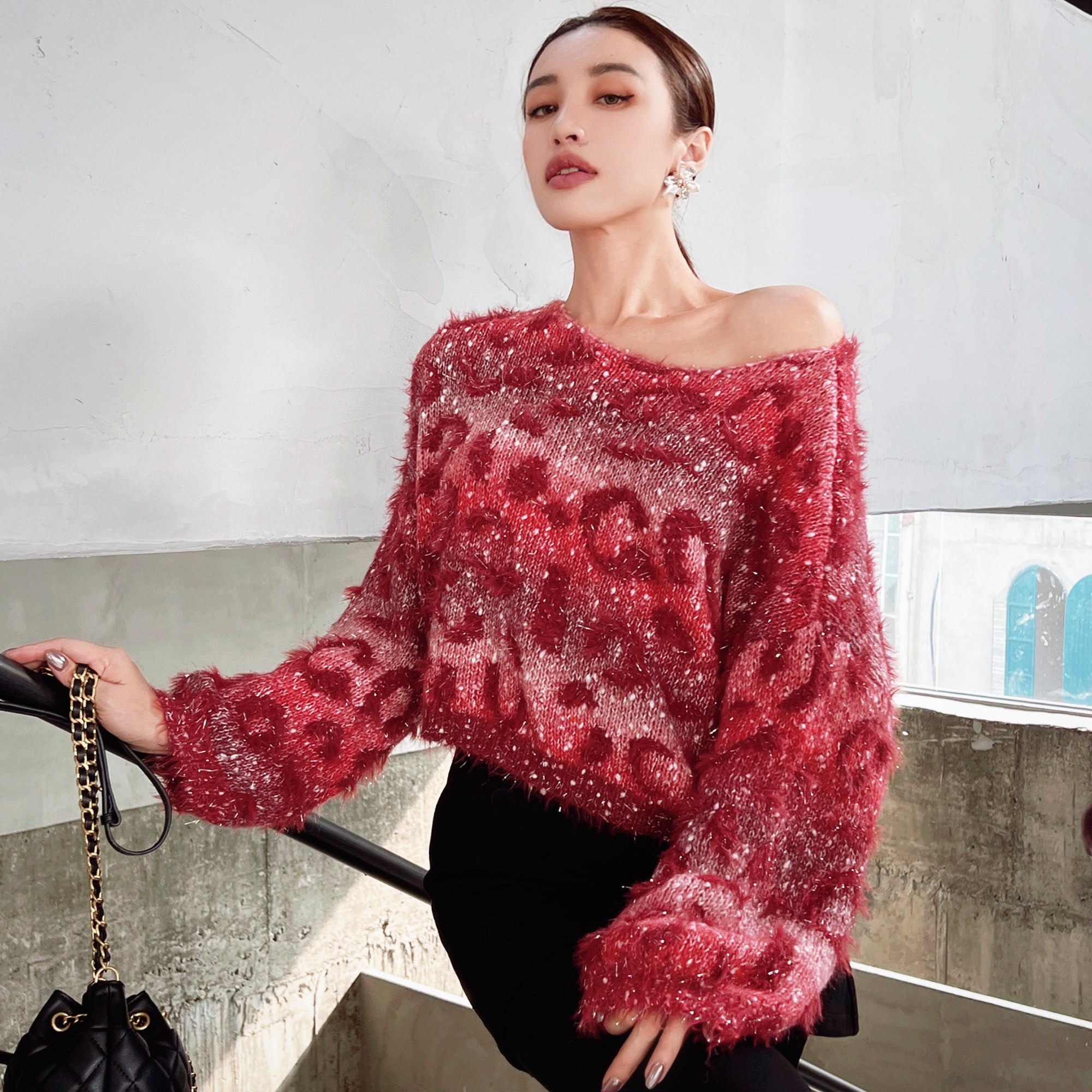 A woman wearing a red, sparkly sweater stands in front of a white wall, holding a black purse.