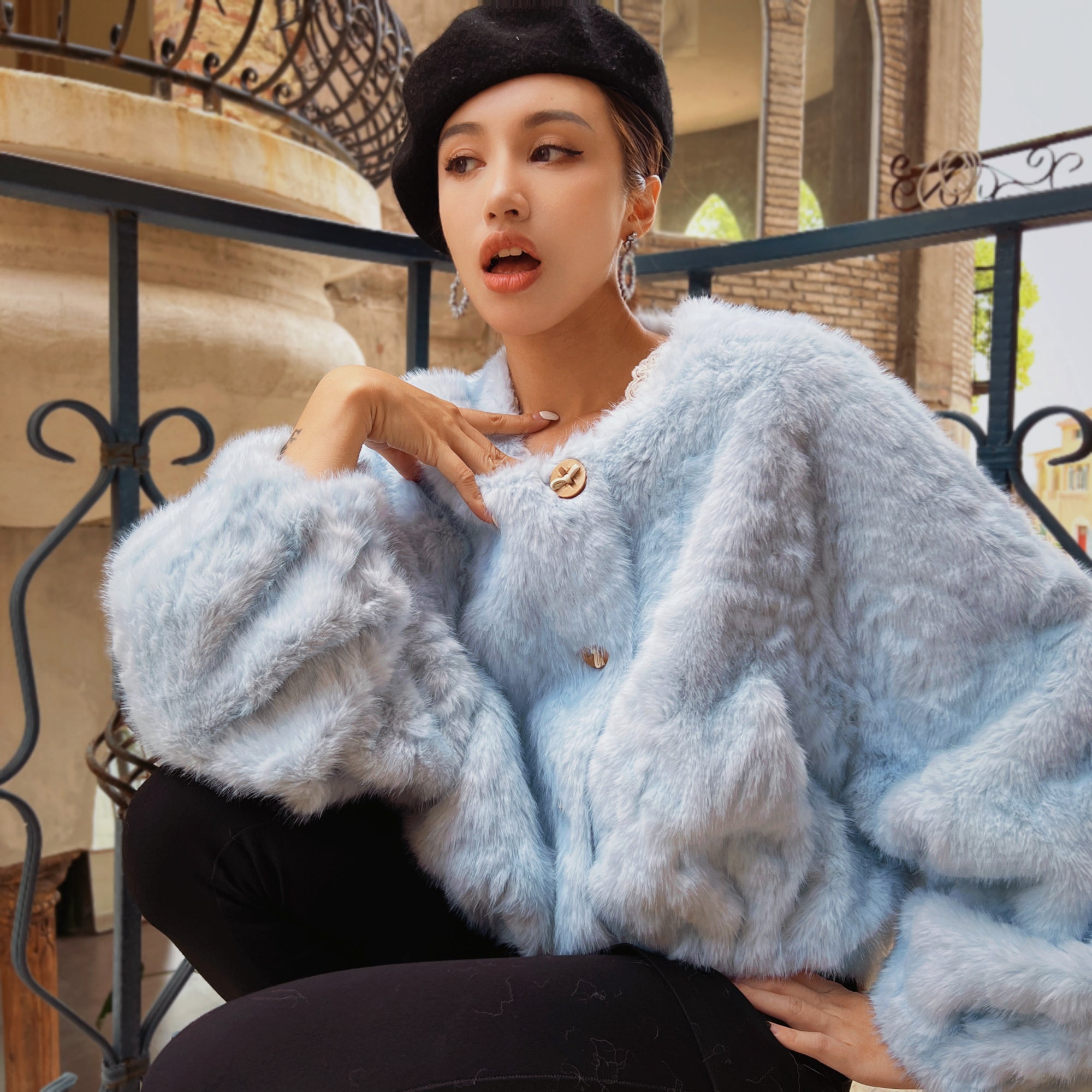 A woman wearing a light blue fur coat and a black hat is sitting on a bench, looking off to the side with her hand on her chin.