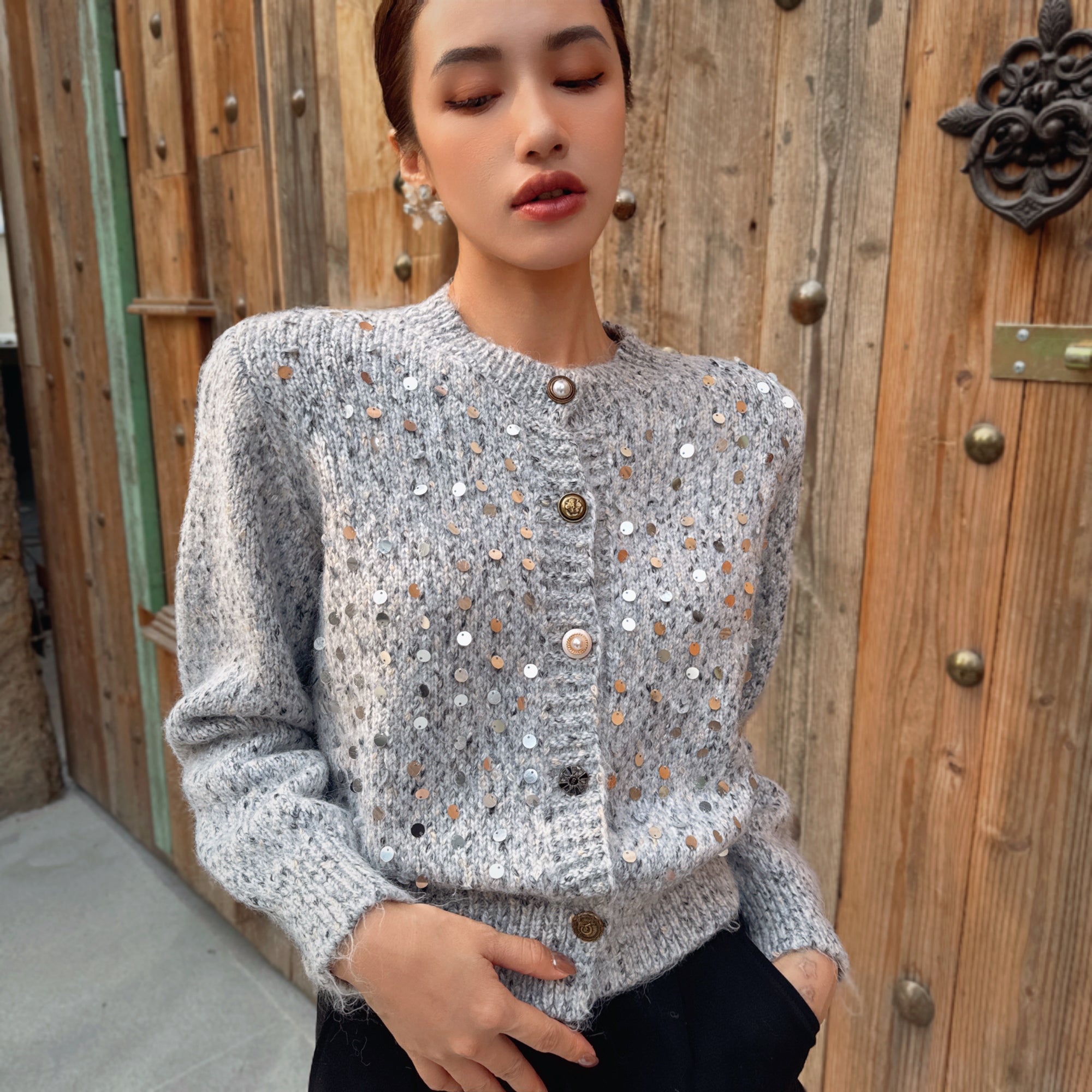 A woman wearing a gray sweater with silver buttons and a patterned design stands in front of a wooden door.