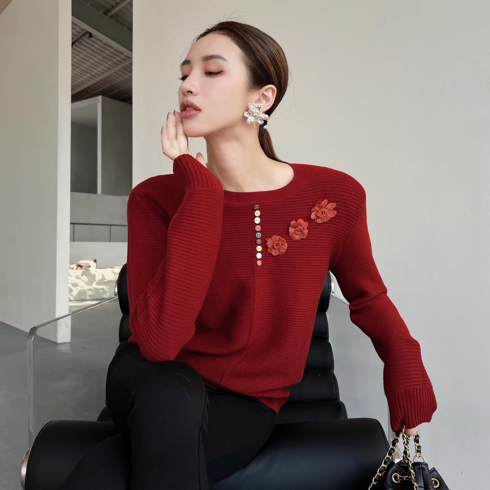 A woman wearing a red sweater with flower embellishments is sitting in a chair, holding a purse and appearing to be in a contemplative or relaxed state.