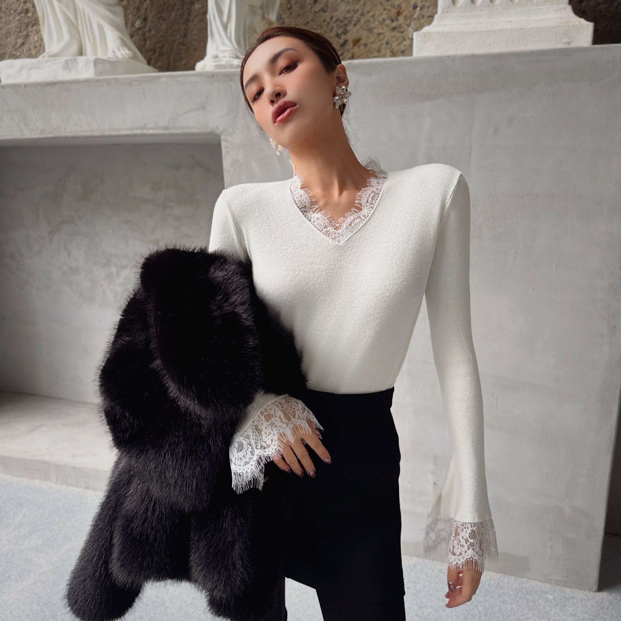 A woman wearing a white top with lace trim and a black fur coat stands in front of a stone wall.