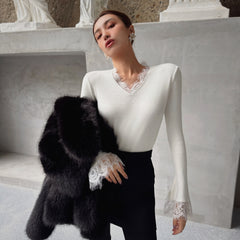 A woman wearing a white top with lace trim and a black fur coat stands in front of a stone wall.