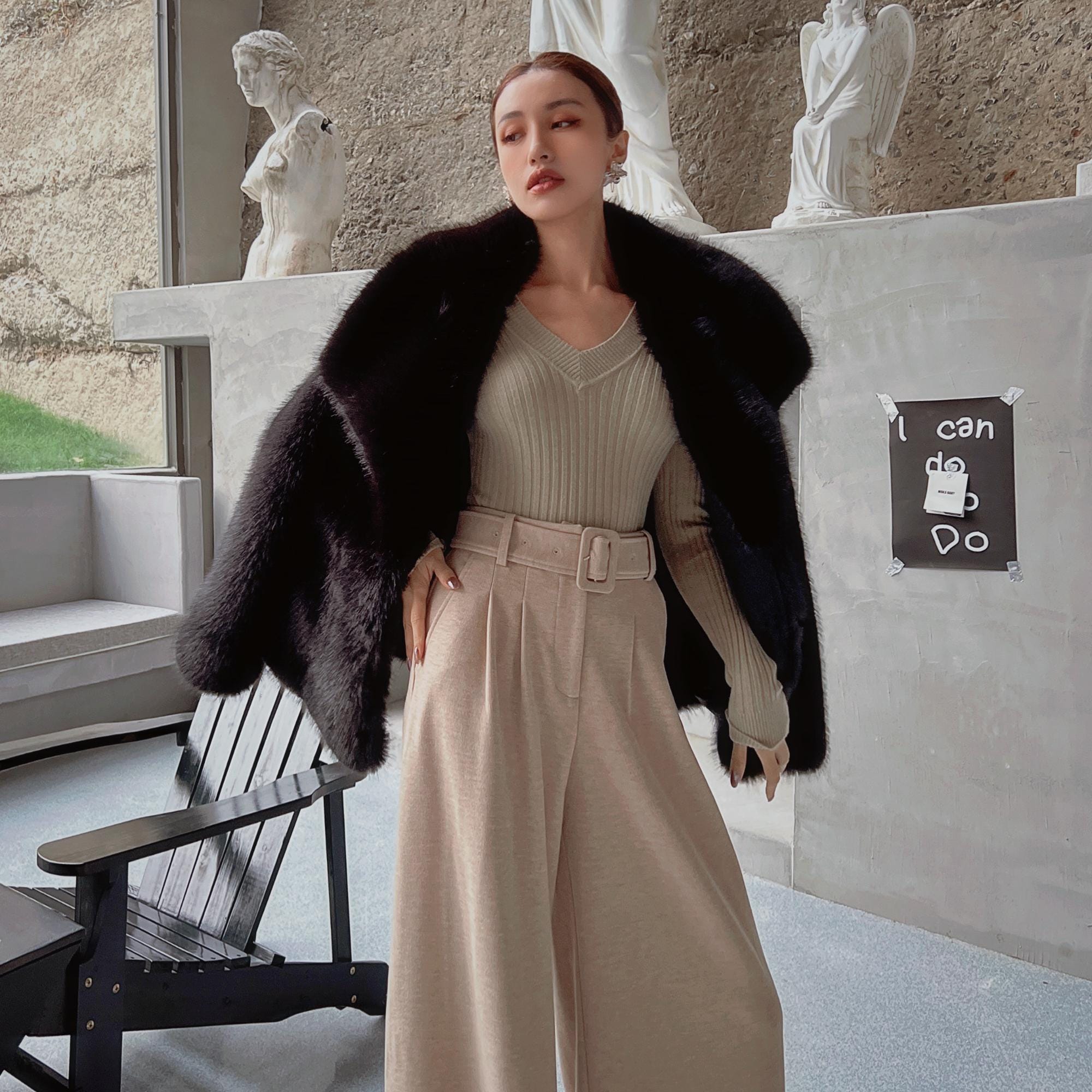 A woman wearing a black fur coat, beige sweater, and wide-leg pants stands in an indoor setting with a stone wall and statues in the background.