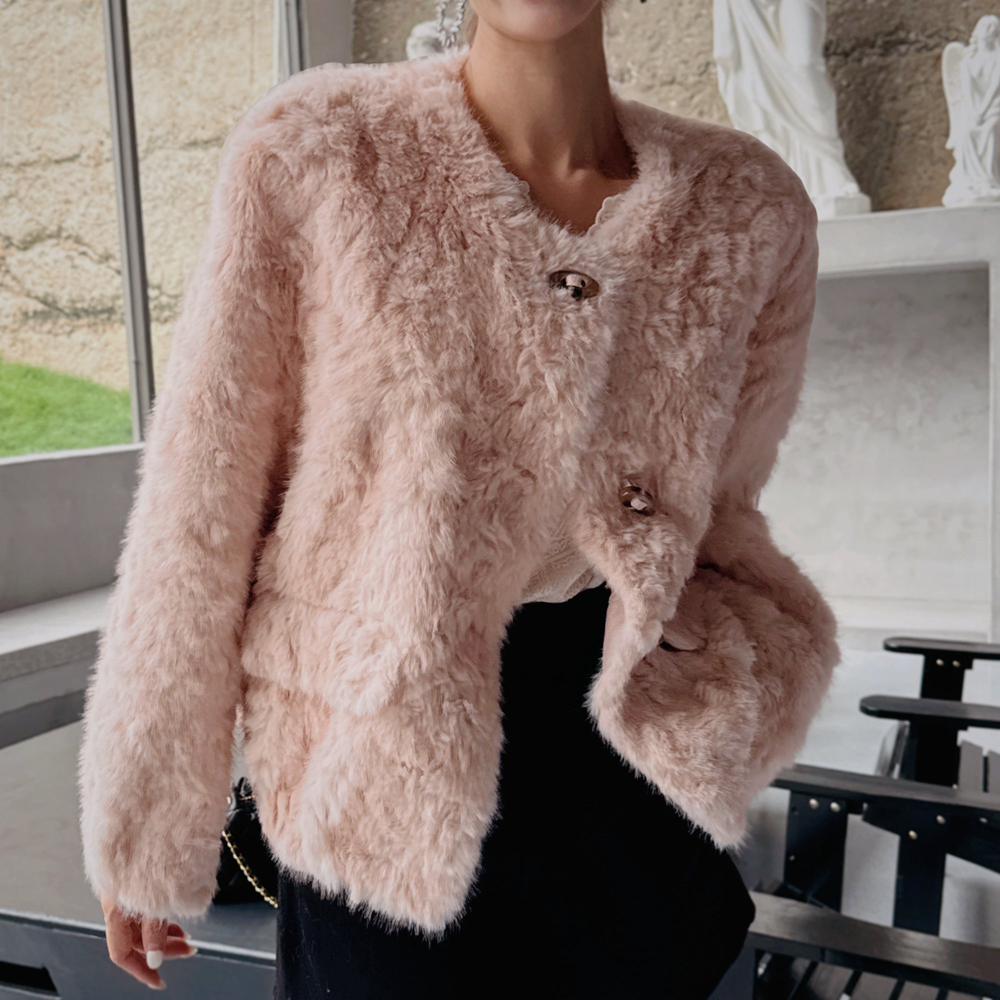 A woman wearing a pink furry coat stands in a room with a window and a statue visible in the background.