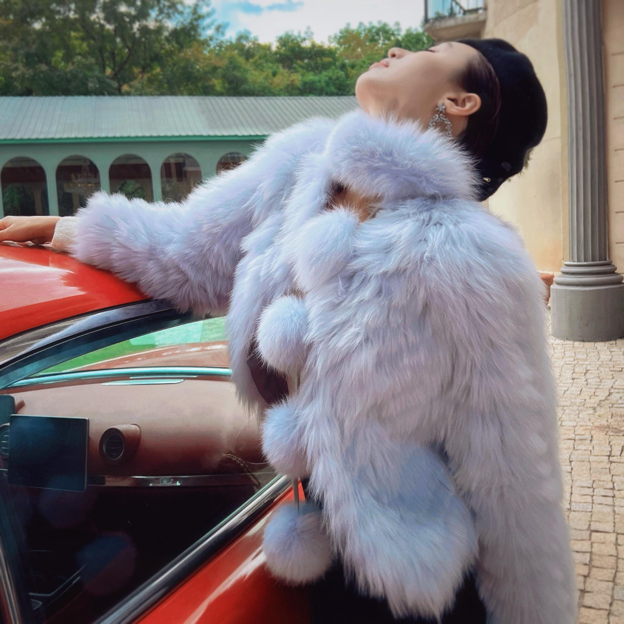 A person wearing a white fur coat stands next to a red car, with their head tilted back and eyes closed, appearing to be in a relaxed and contemplative state.