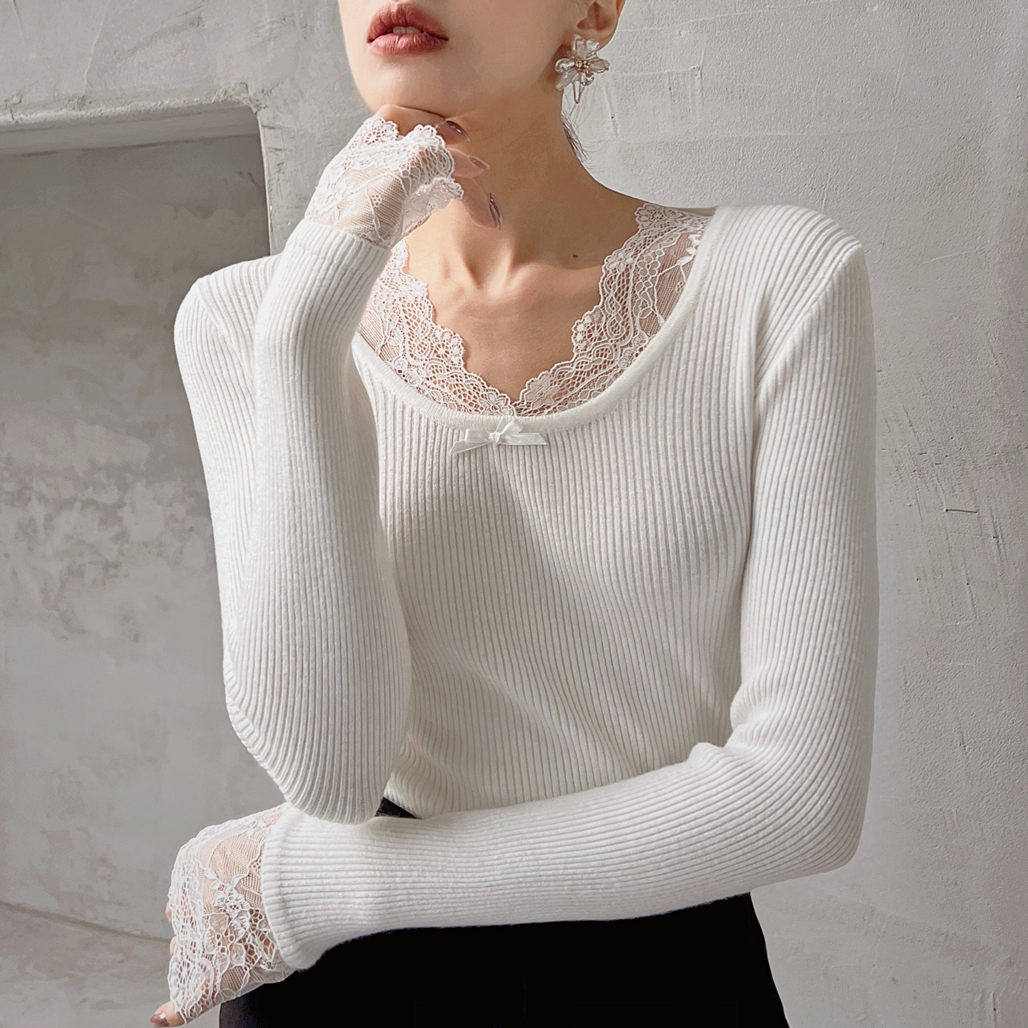 A woman wearing a white lace top and a black skirt, posing with her hand on her chin.