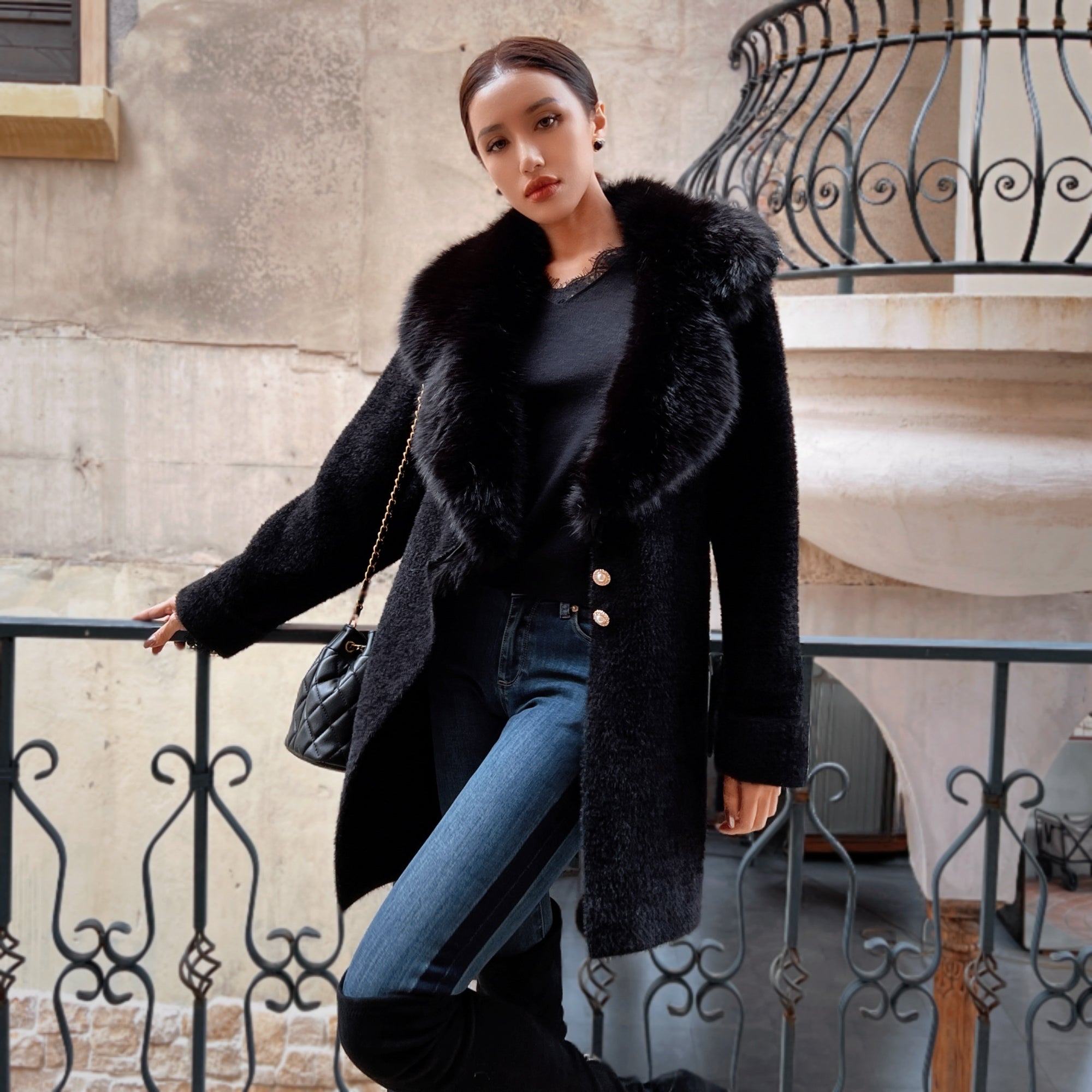 A woman wearing a black fur coat, black top, and blue jeans stands in front of a wrought iron fence, leaning against it with her hand on the railing.