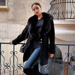 A woman wearing a black fur coat, black top, and blue jeans stands in front of a wrought iron fence, leaning against it with her hand on the railing.
