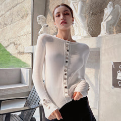 A woman wearing a white sweater with buttons poses in an indoor setting with a stone wall and statues in the background.