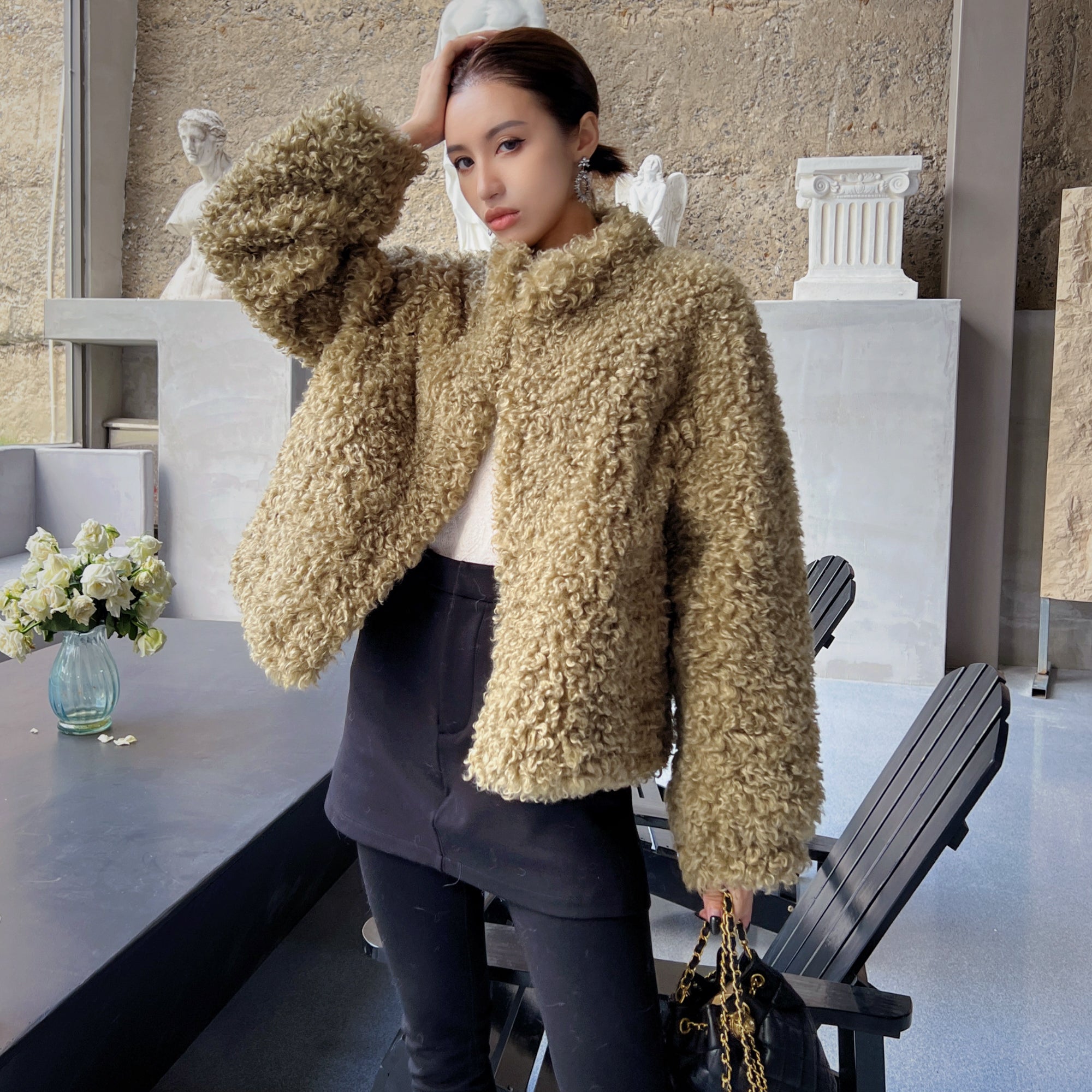 A woman wearing a beige fur coat stands in a room with a stone wall and a vase of flowers on a table.
