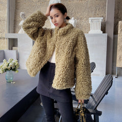 A woman wearing a beige fur coat stands in a room with a stone wall and a vase of flowers on a table.