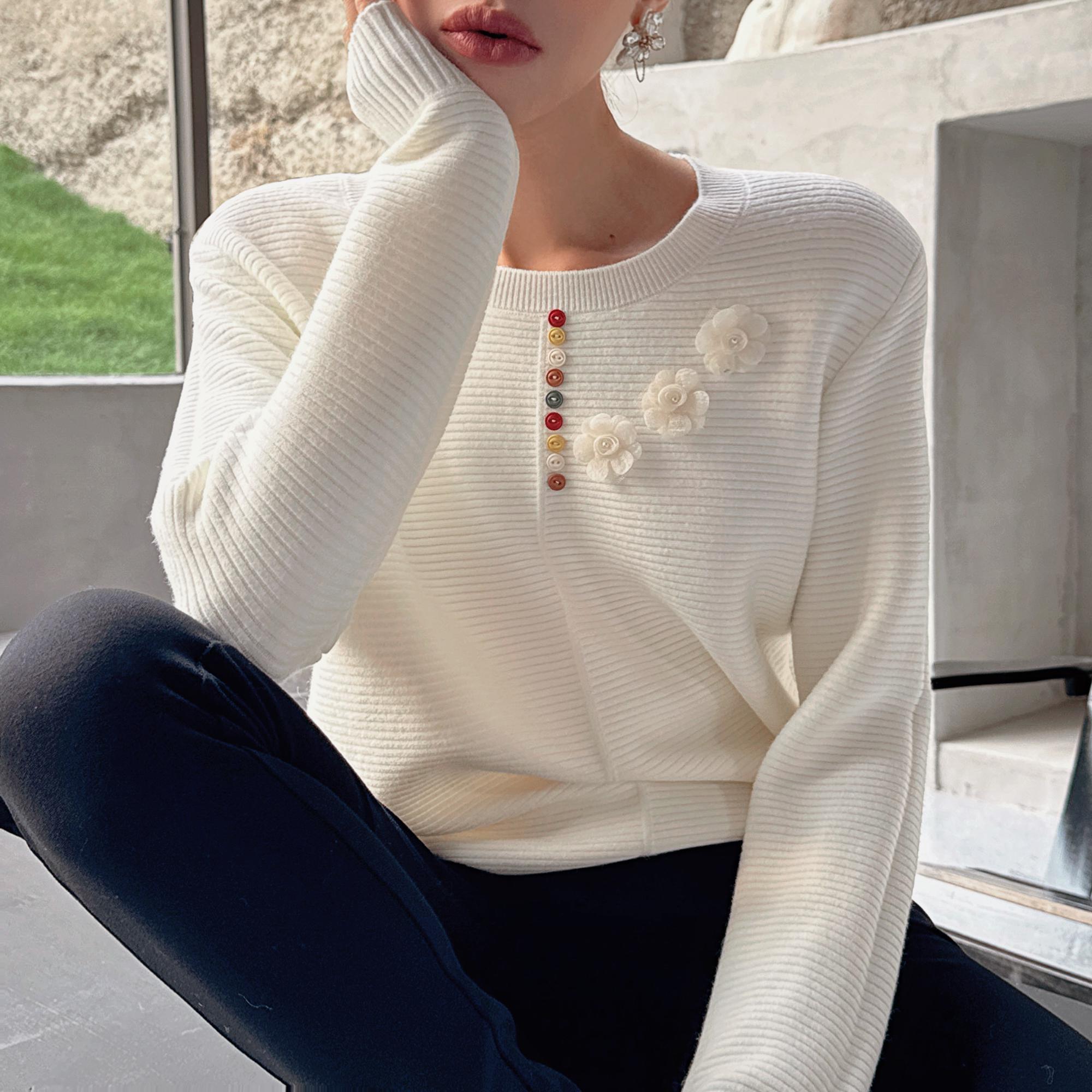 A woman wearing a white sweater with colorful buttons and a flower design, sitting with her hand on her chin.