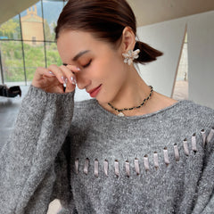 A woman wearing a gray sweater with a floral pattern and a necklace is shown in a close-up shot, with her eyes closed and her hand touching her face.