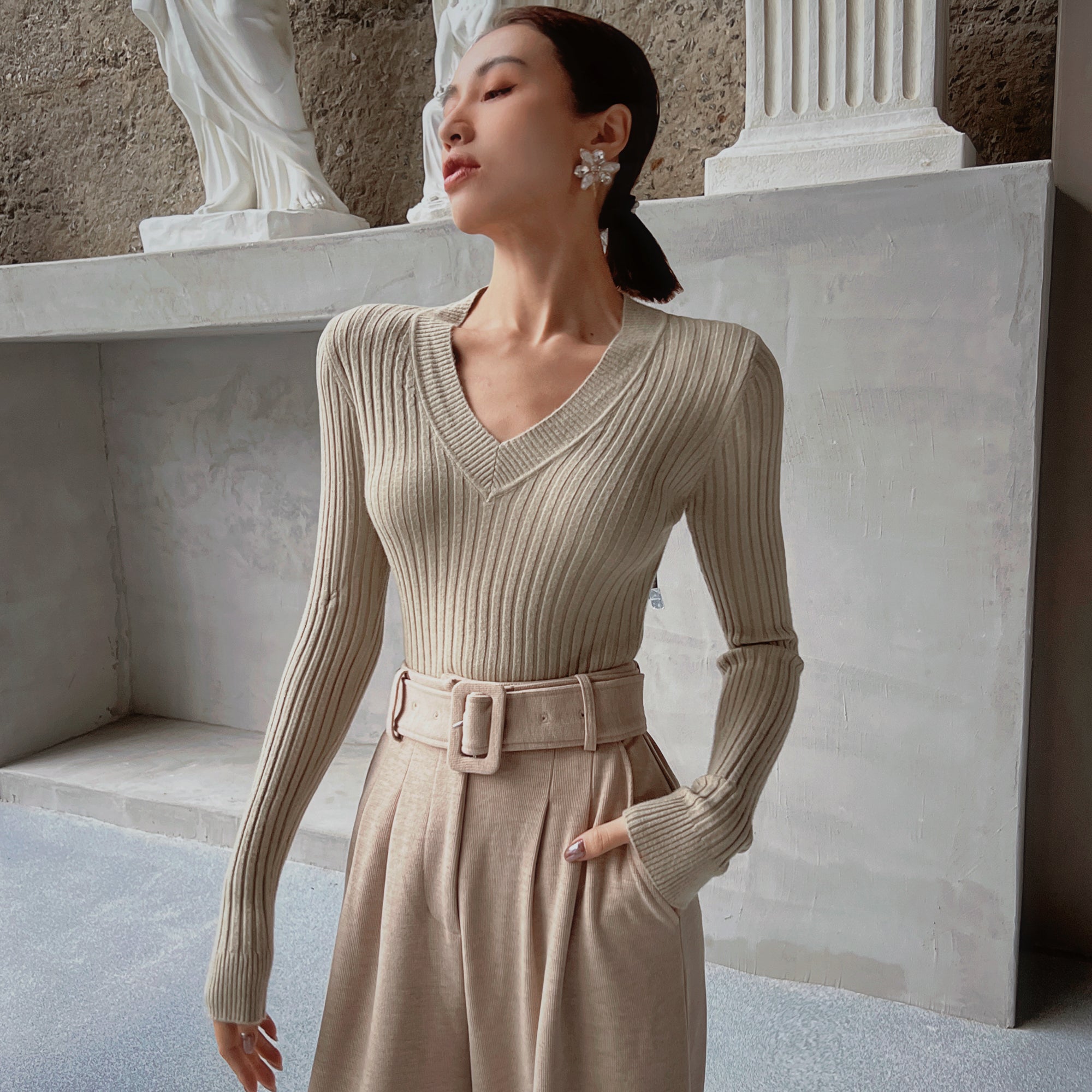 A woman wearing a beige sweater and a belt, standing in front of a stone wall with a statue on top.