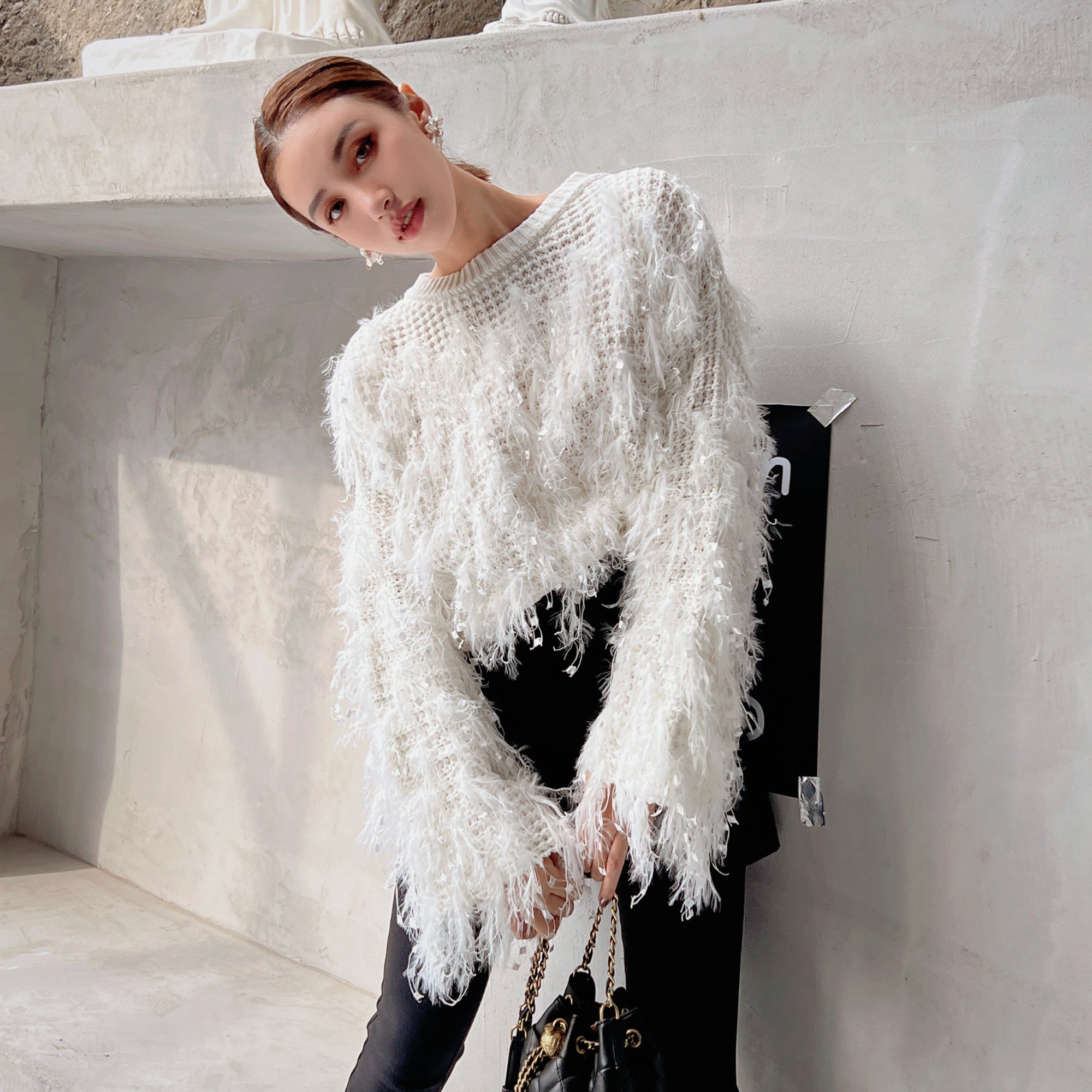 A woman wearing a white fuzzy sweater and holding a black purse is posing against a white wall.