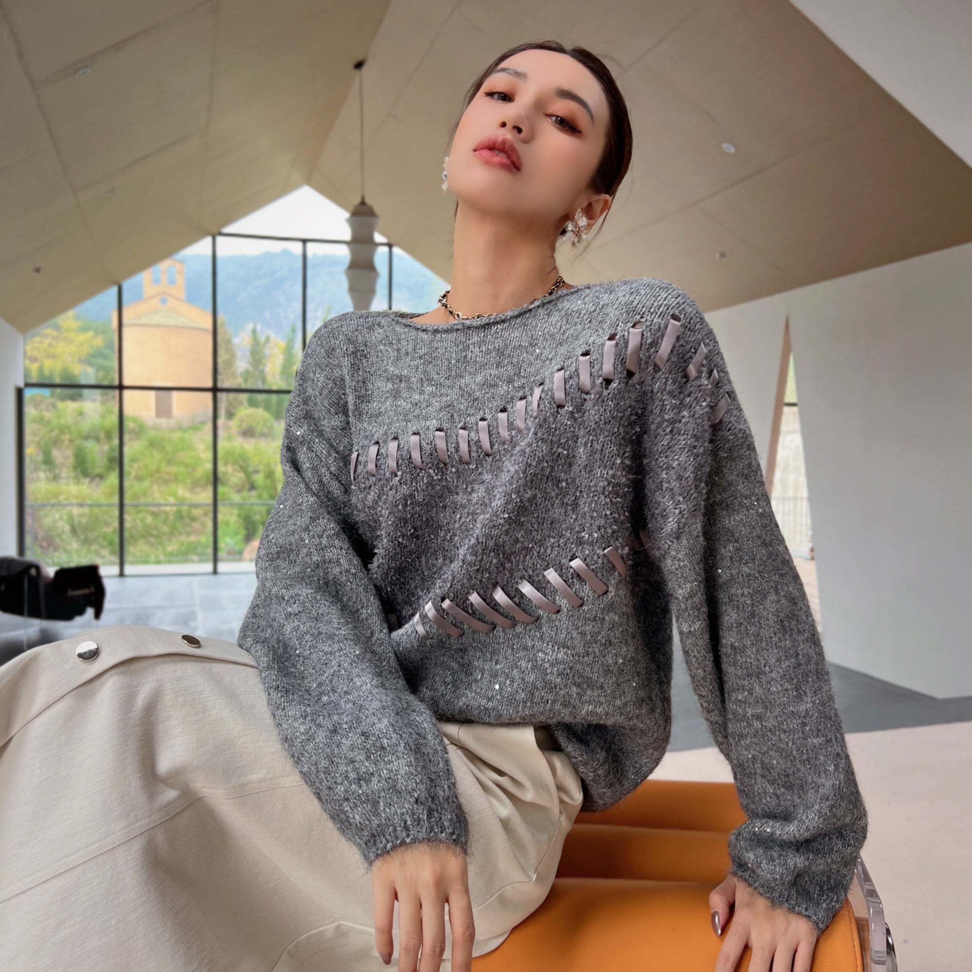 A woman wearing a gray sweater with silver stitching and a beige skirt is sitting on an orange bench in a room with large windows overlooking a mountainous landscape.