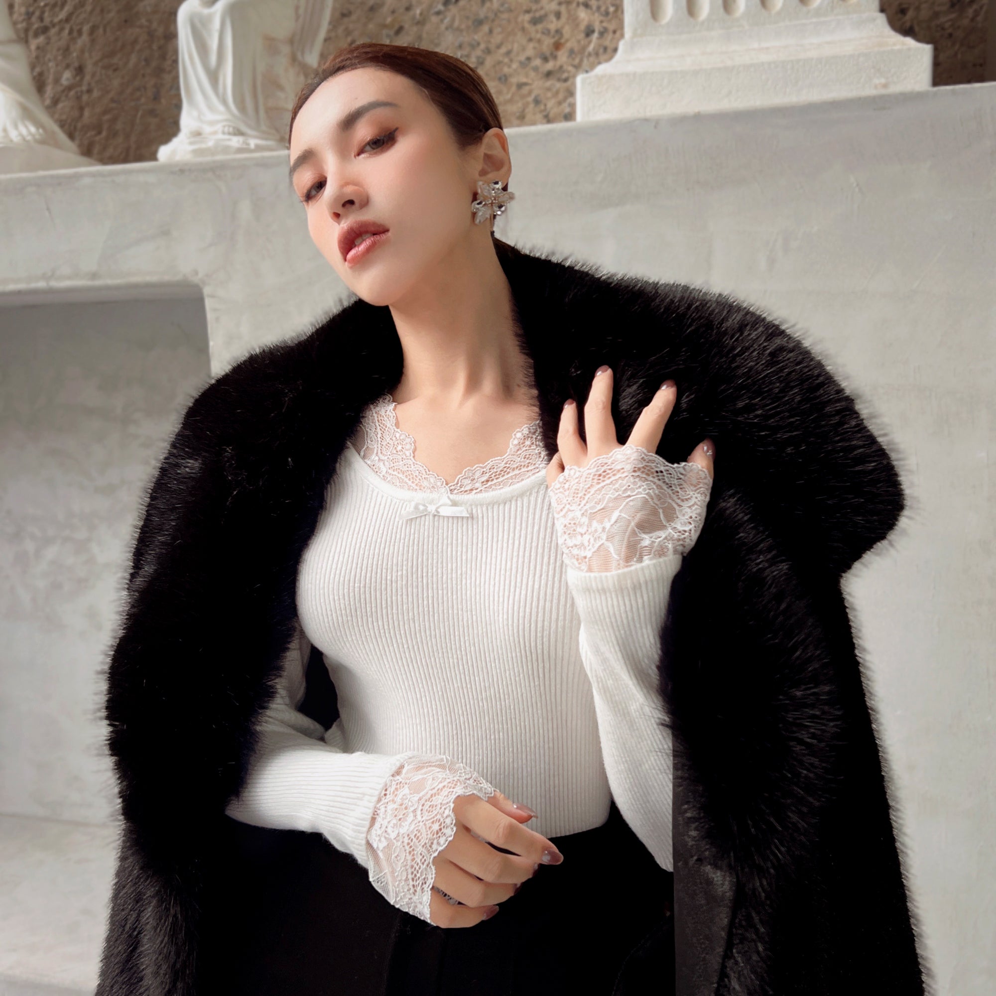 A woman wearing a white lace top and a black fur coat stands in front of a white wall, with her hand resting on her chin.