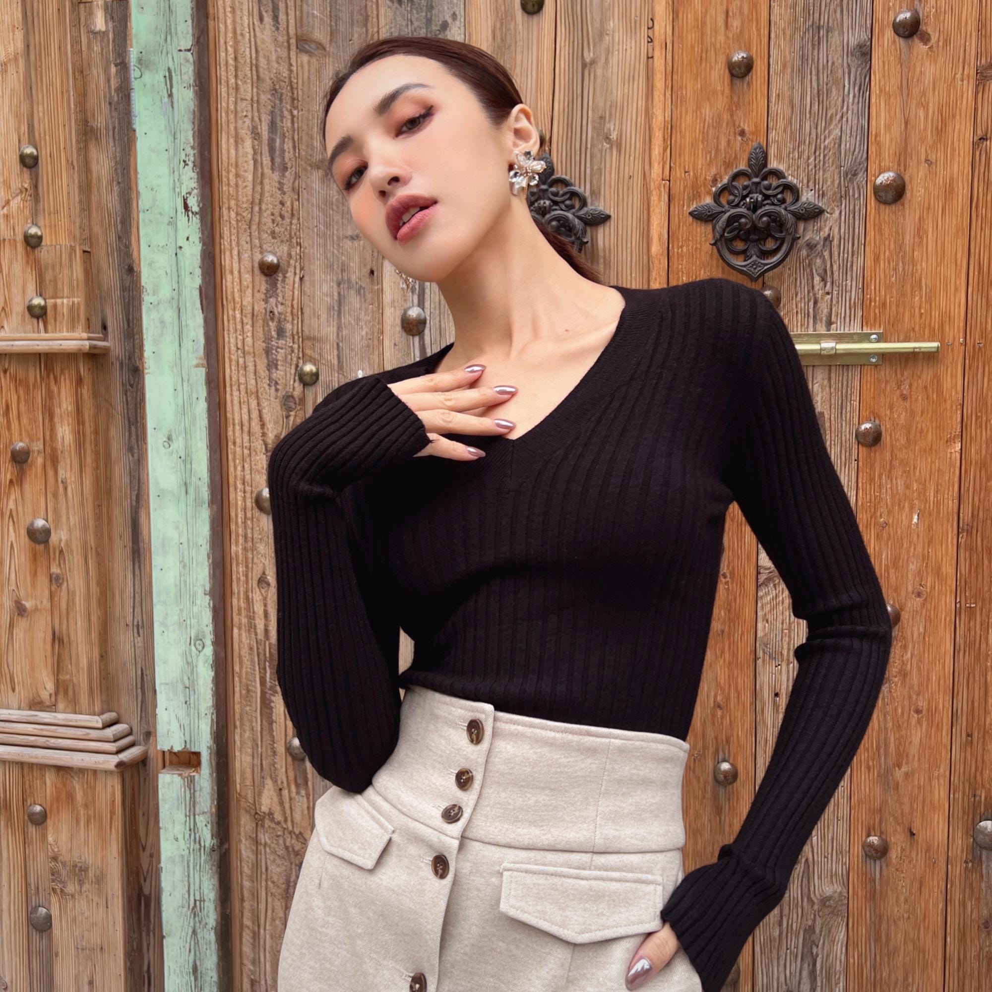 A woman wearing a black sweater and a beige skirt stands in front of a wooden door with a floral design.